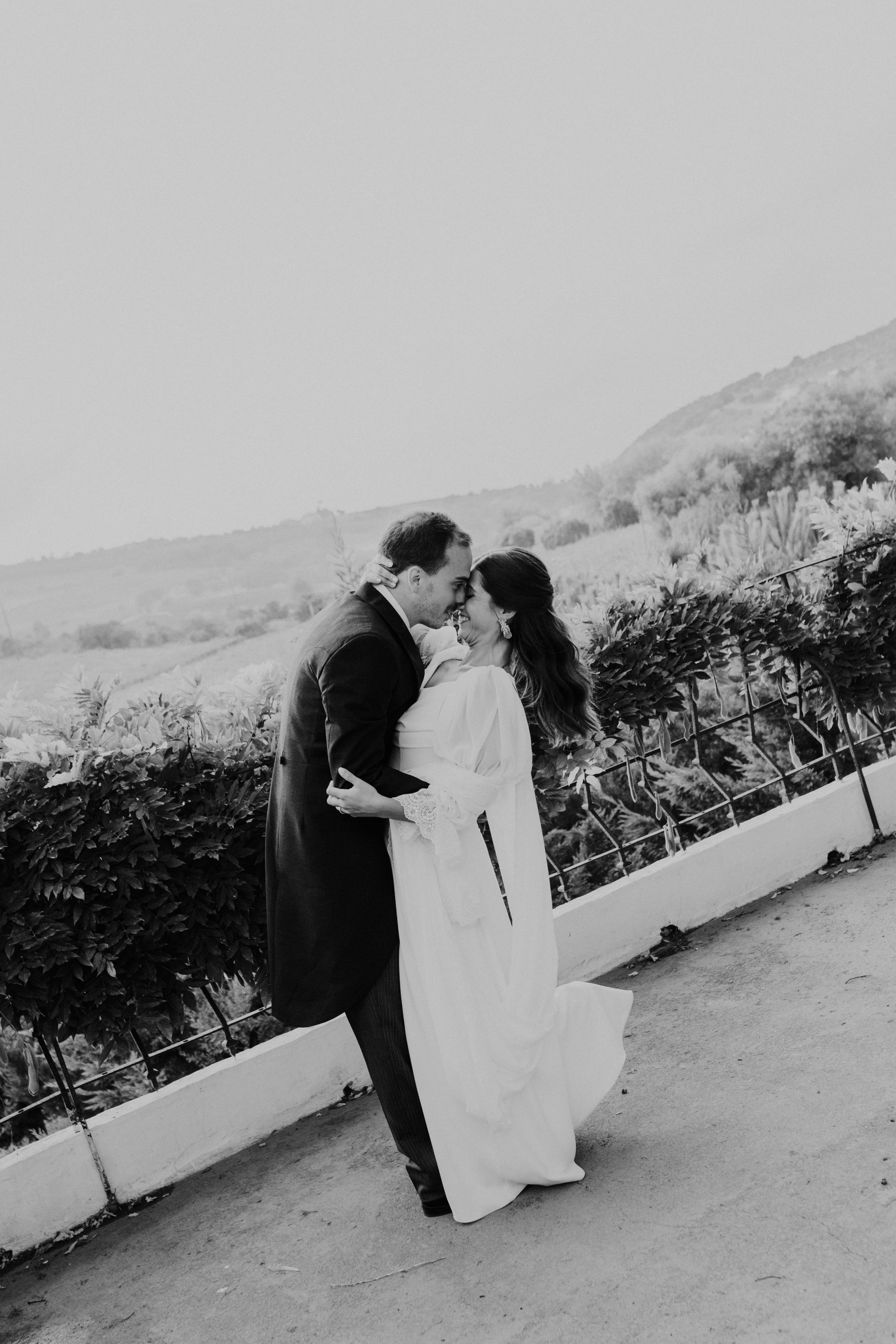 Black and white photo of a bride and groom embracing and smiling in front of a scenic countryside backdrop.