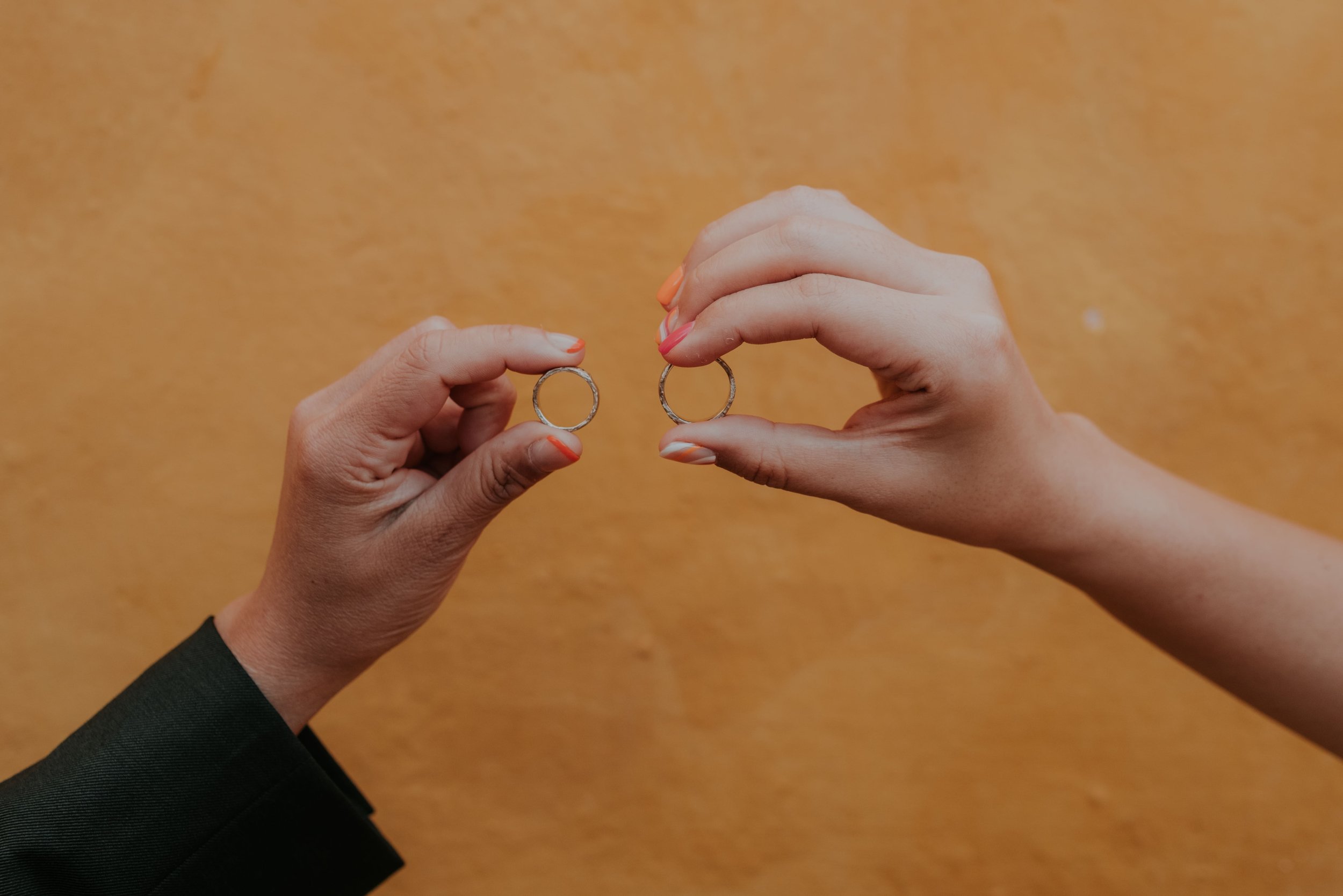 Two hands holding wedding rings in front of a textured mustard yellow wall, featuring vibrant wedding nail designs. Captured by Carlos Mendes Photography.