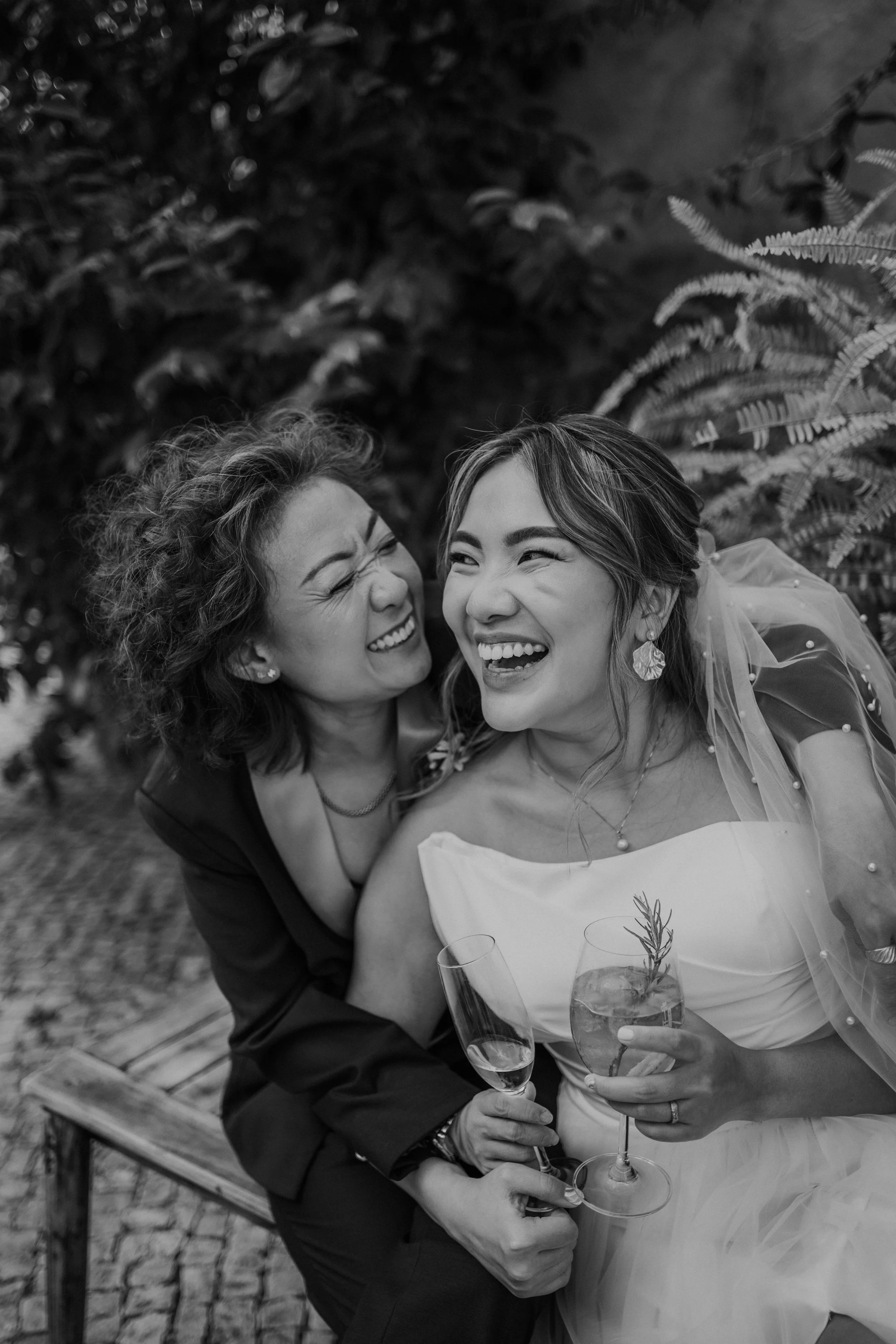 Black and white close-up of two joyful brides sitting together, laughing and holding wedding drinks, captured with emotion and intimacy by Carlos Mendes Photography.