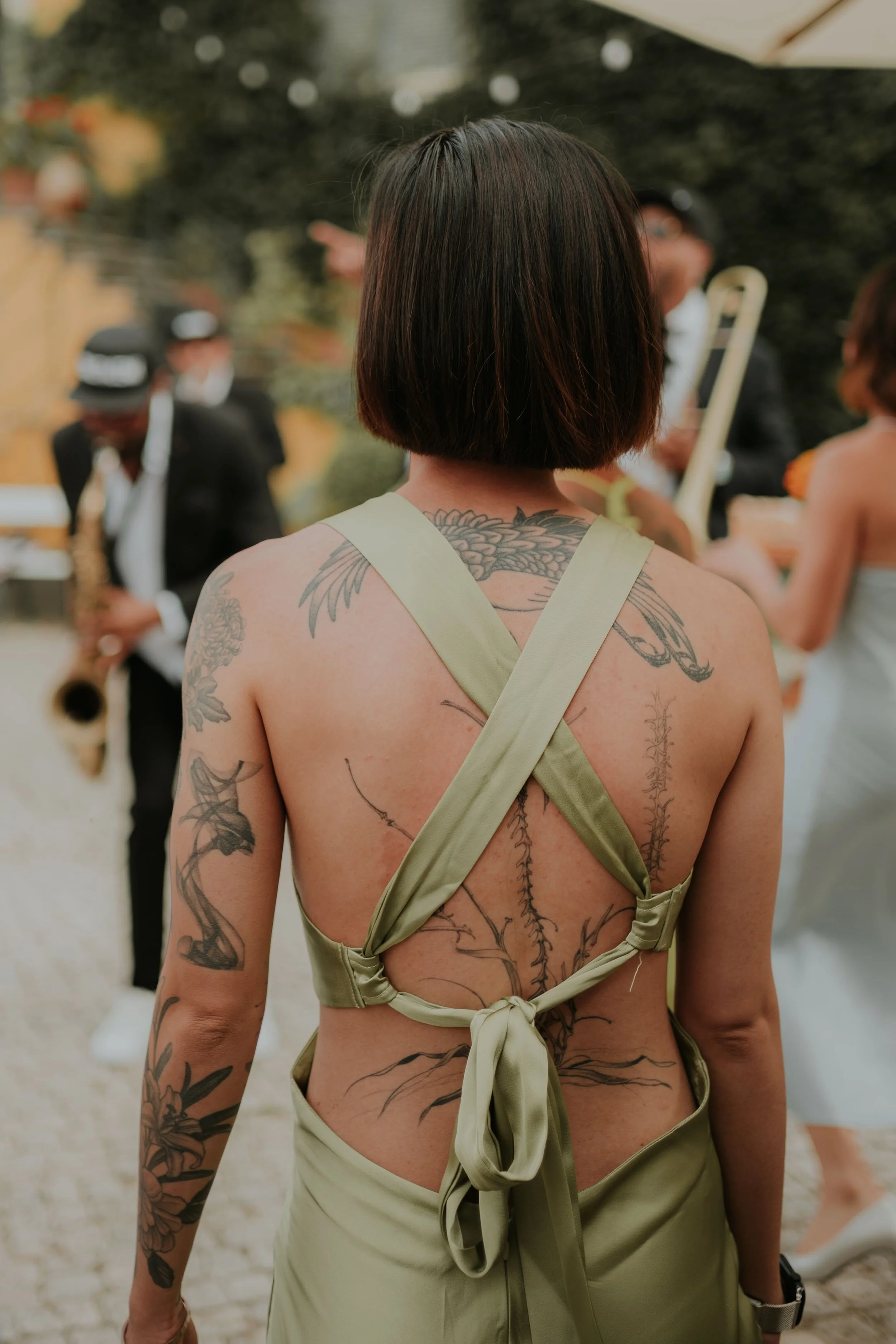 Wedding guest with intricate botanical tattoos wearing a sage green backless dress, watching live jazz musicians during the cocktail hour. Captured by Carlos Mendes Photography.
