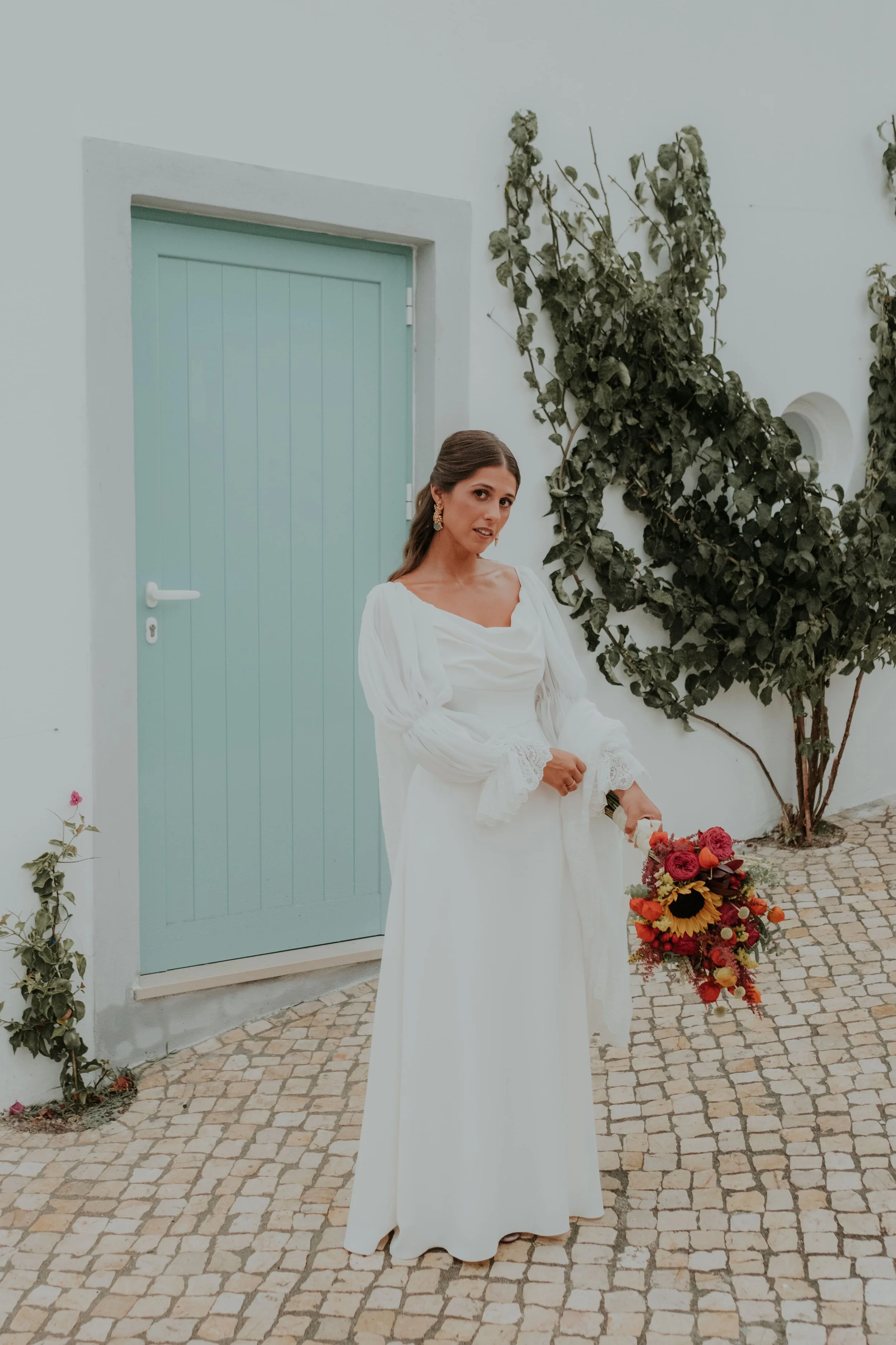 Bride in a white dress holding a colorful bouquet, standing in front of a pastel green door and white wall with vines.