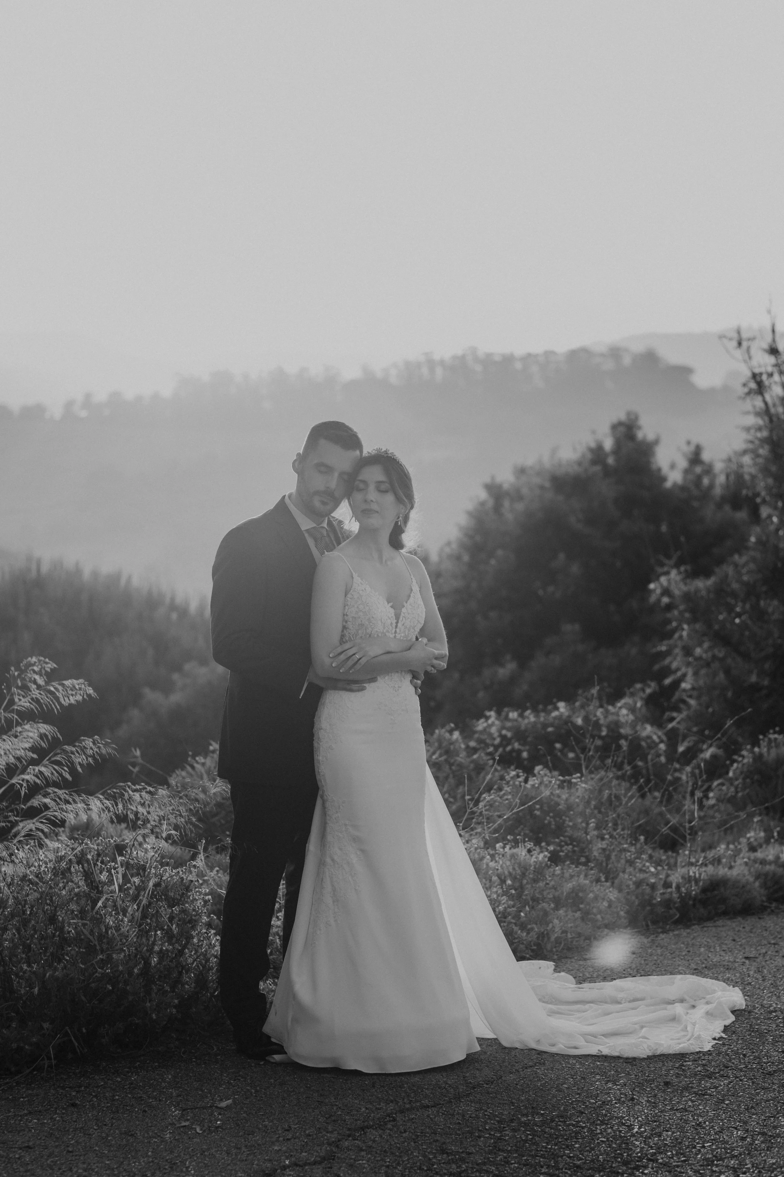Black and white portrait of a bride and groom embracing with eyes closed, surrounded by soft natural light and lush countryside landscape, captured by Carlos Mendes Photography.