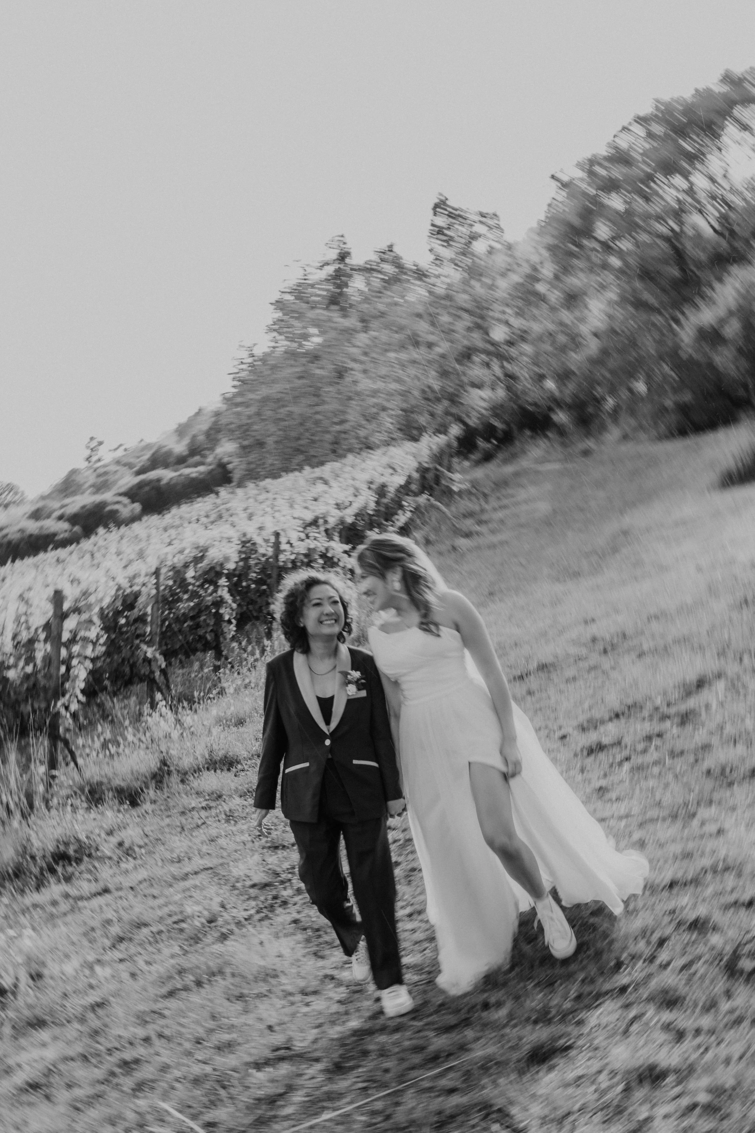 Two brides walking hand in hand through a vineyard during their same-sex wedding day, smiling and enjoying the moment in nature, captured by Carlos Mendes Photography.