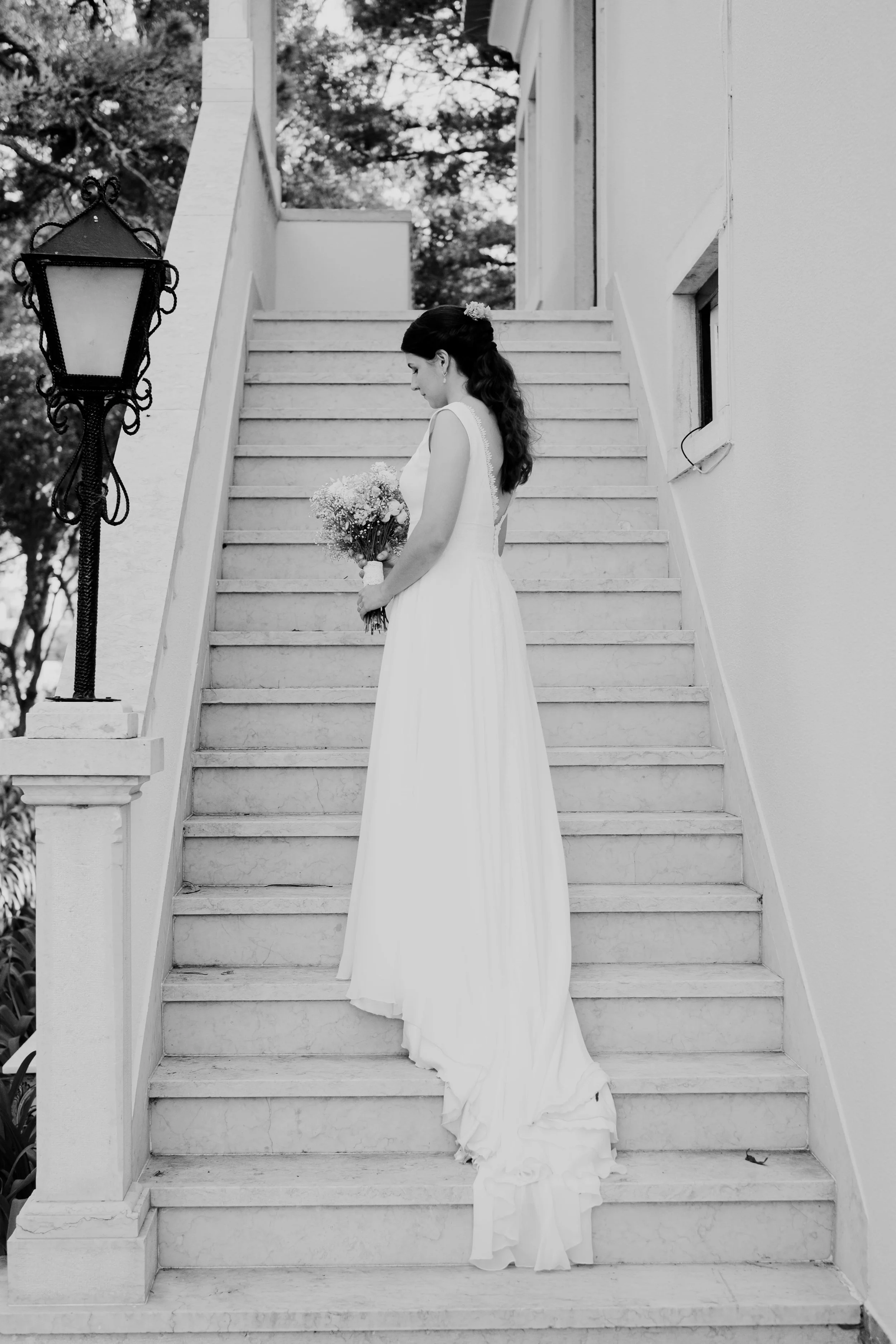 Black and white photo of a bride standing gracefully on a stone staircase, holding a bouquet and looking down with a serene expression, captured by Carlos Mendes Photography.