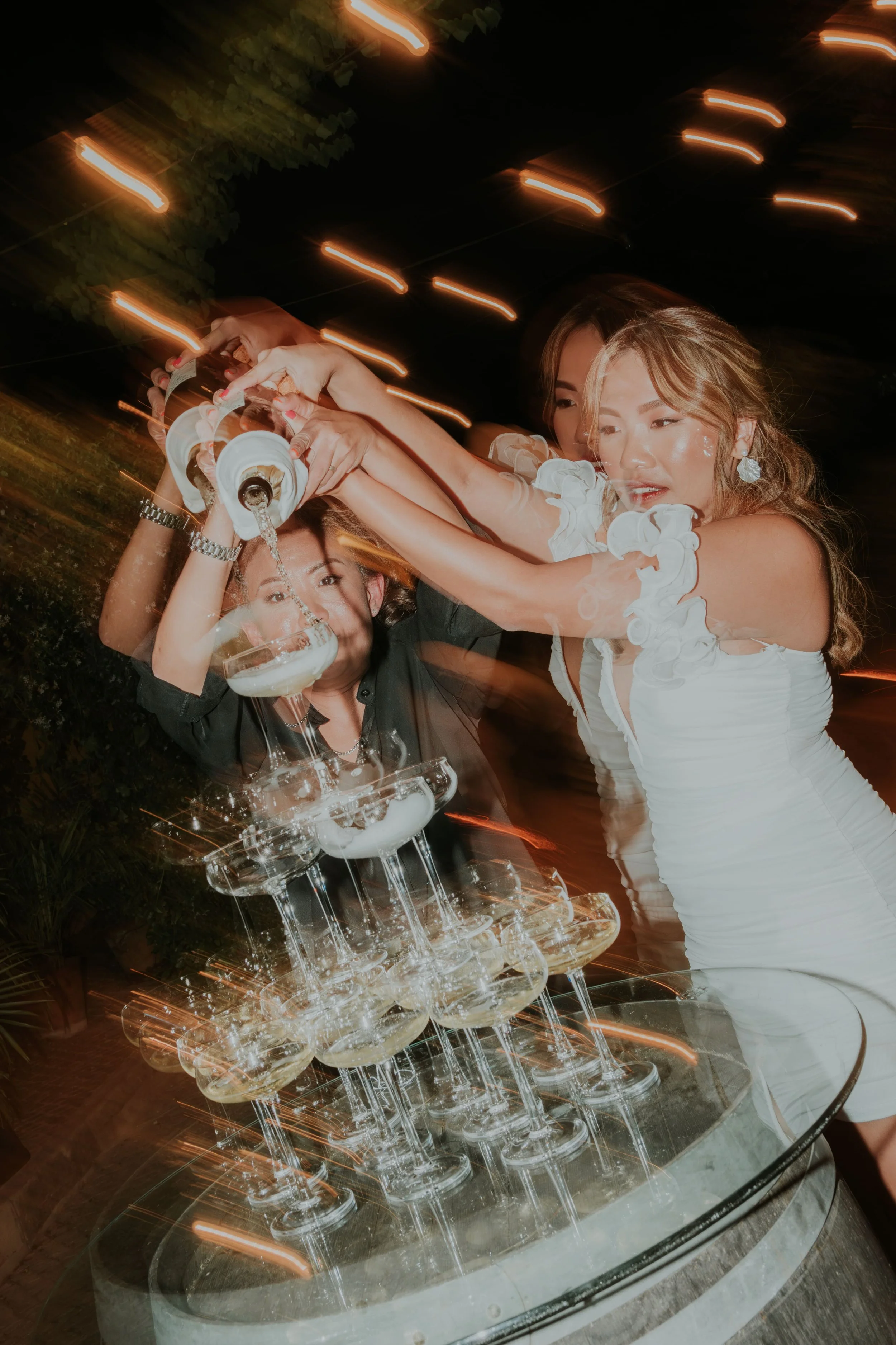 Energetic photo of a bride and her friends pouring champagne into a glass tower during a lively wedding party at night, with motion blur and warm string lights, captured by Carlos Mendes Photography