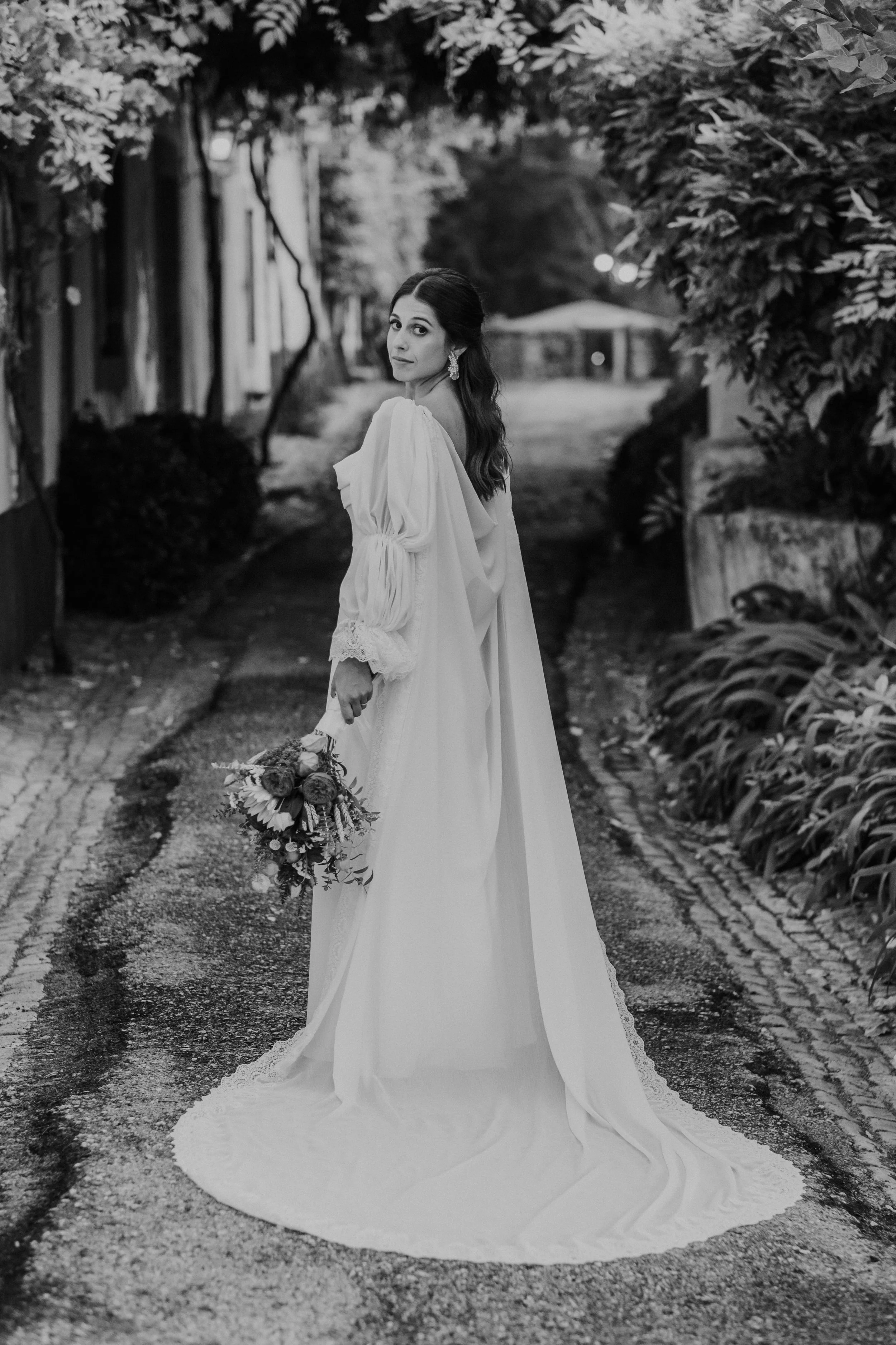 Bride in a long wedding dress looks back while walking down a garden path, holding a vibrant bouquet. Black and white portrait.