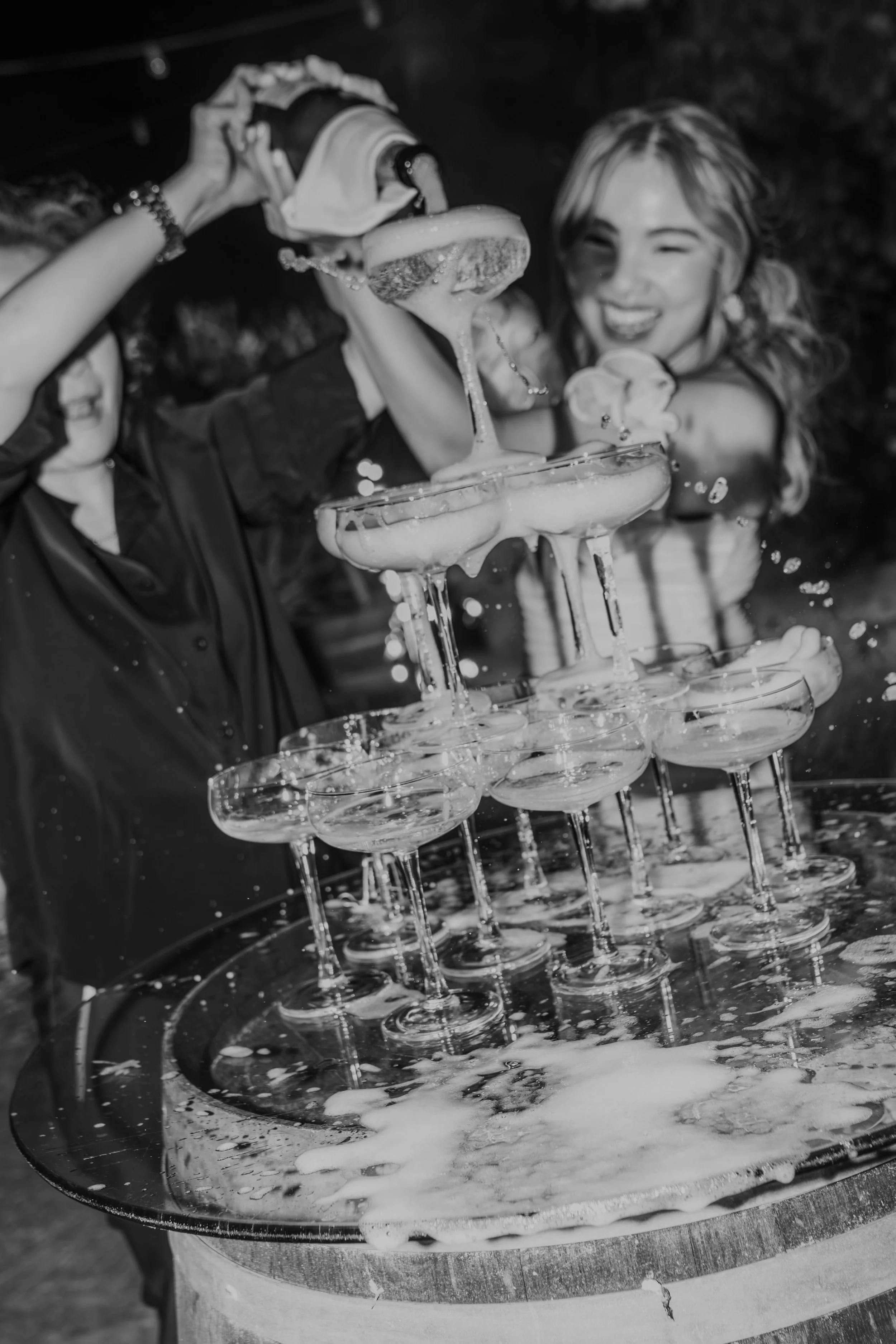 Black and white photo of champagne overflowing in a glass tower as the bride and a guest pour bottles during a lively night wedding celebration, captured by Carlos Mendes Photography.