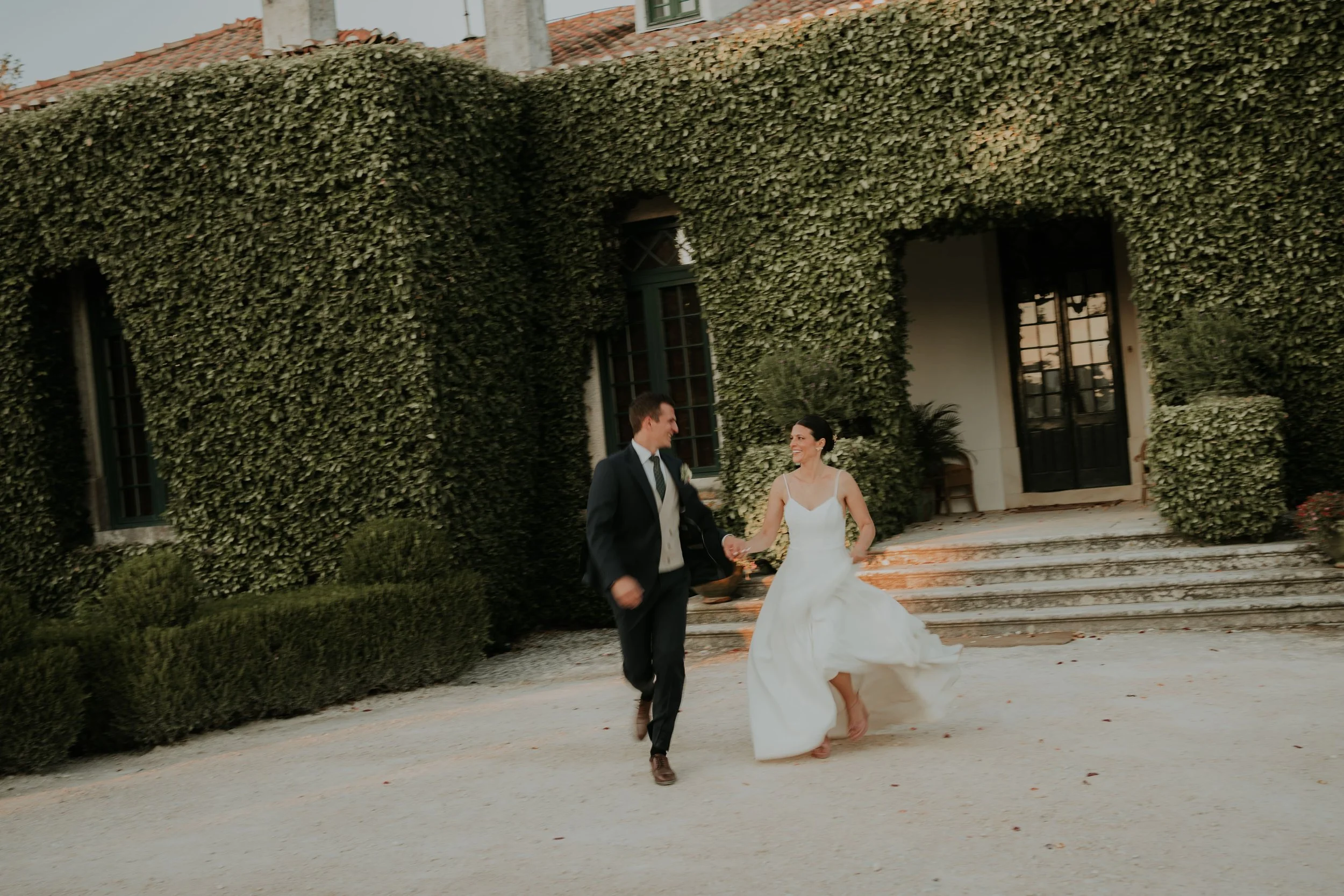 Bride and groom running joyfully in front of a vintage mansion covered in ivy, holding hands and smiling at each other."