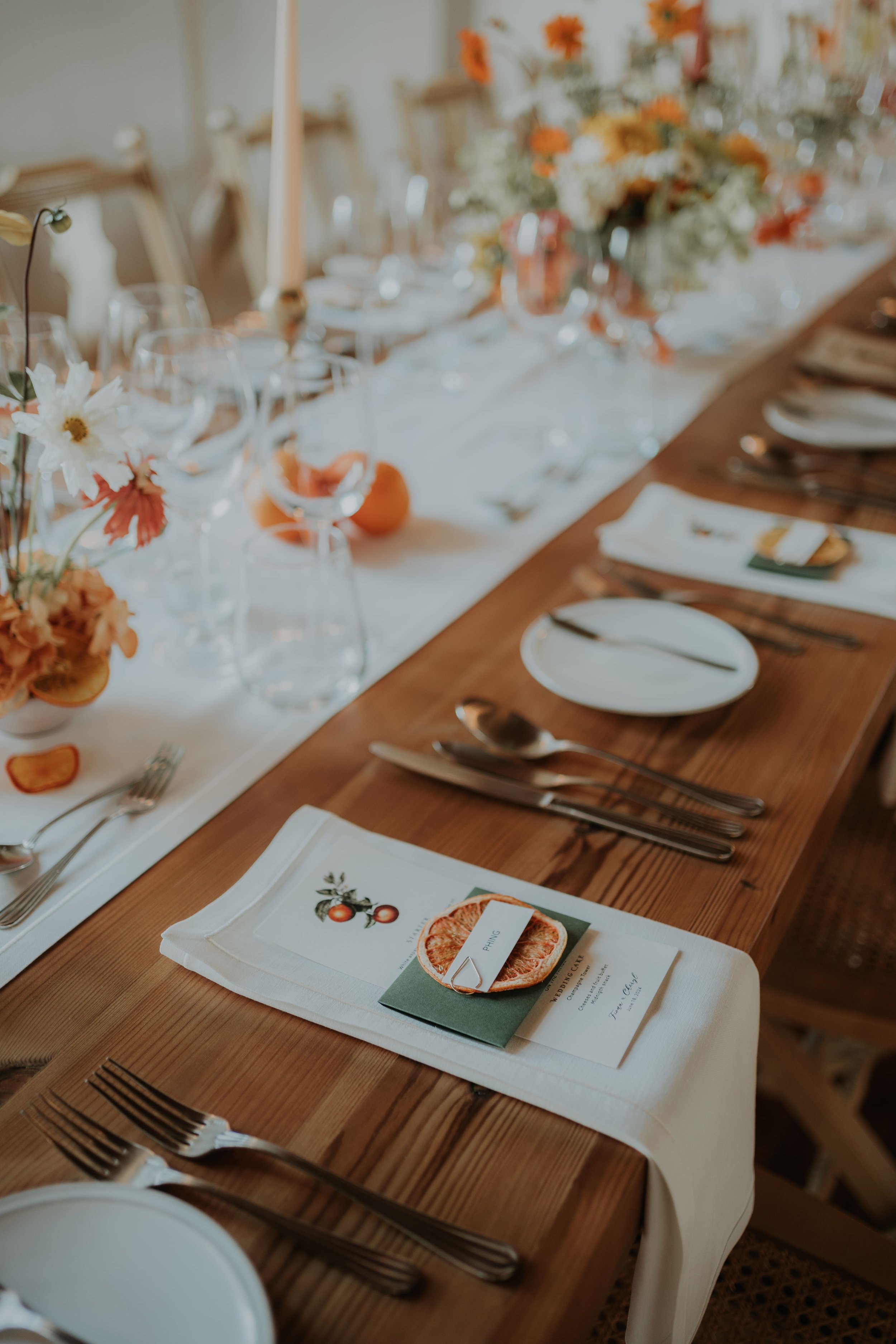 Close-up of a wedding table setup featuring citrus-themed menu cards, dried orange slices, elegant cutlery, and fresh floral centerpieces in shades of white and orange. Styled and captured by Carlos Mendes Photography.