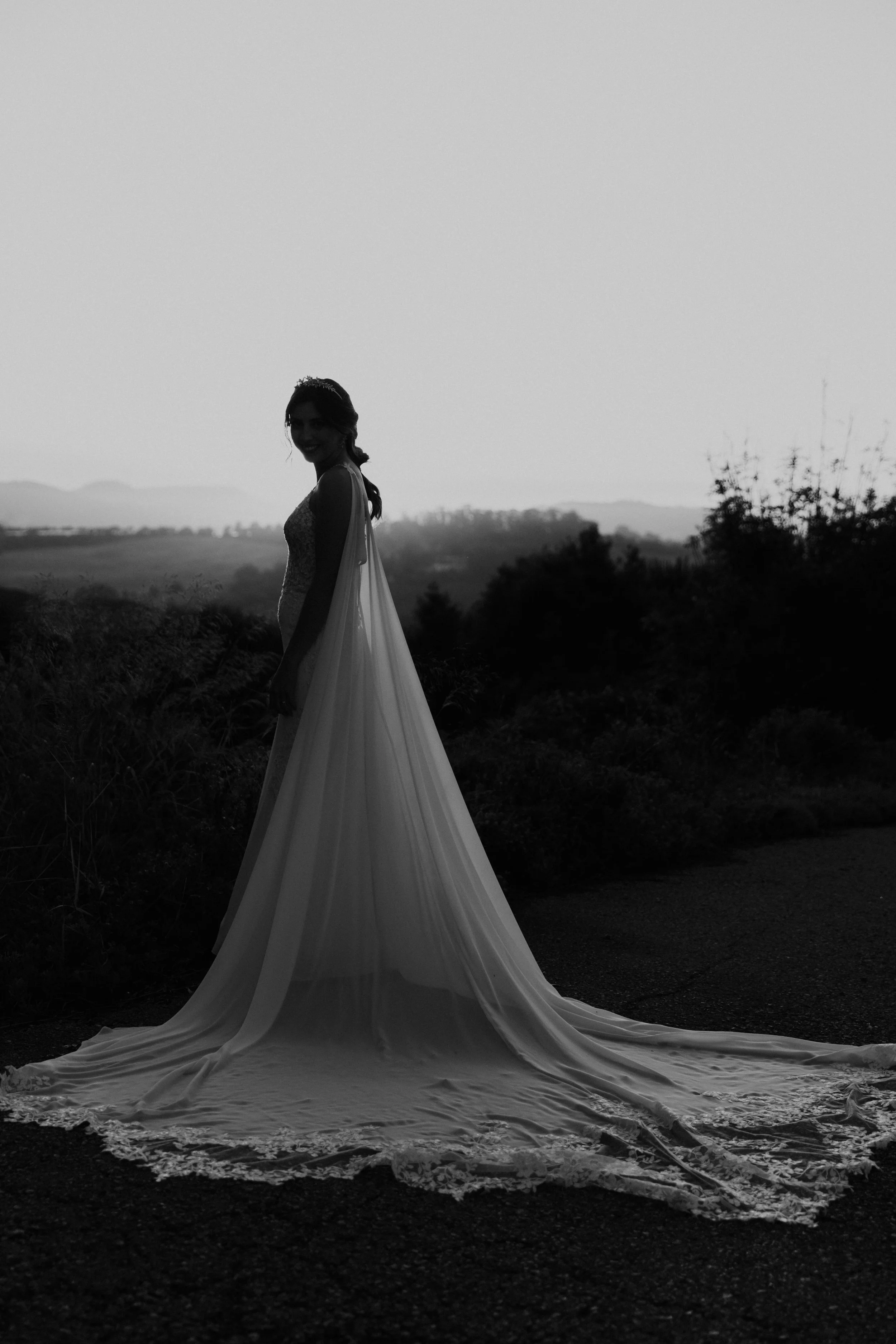 Black and white silhouette of a bride in a long lace-trimmed wedding gown at sunset, standing peacefully in nature, captured by Carlos Mendes Photography.