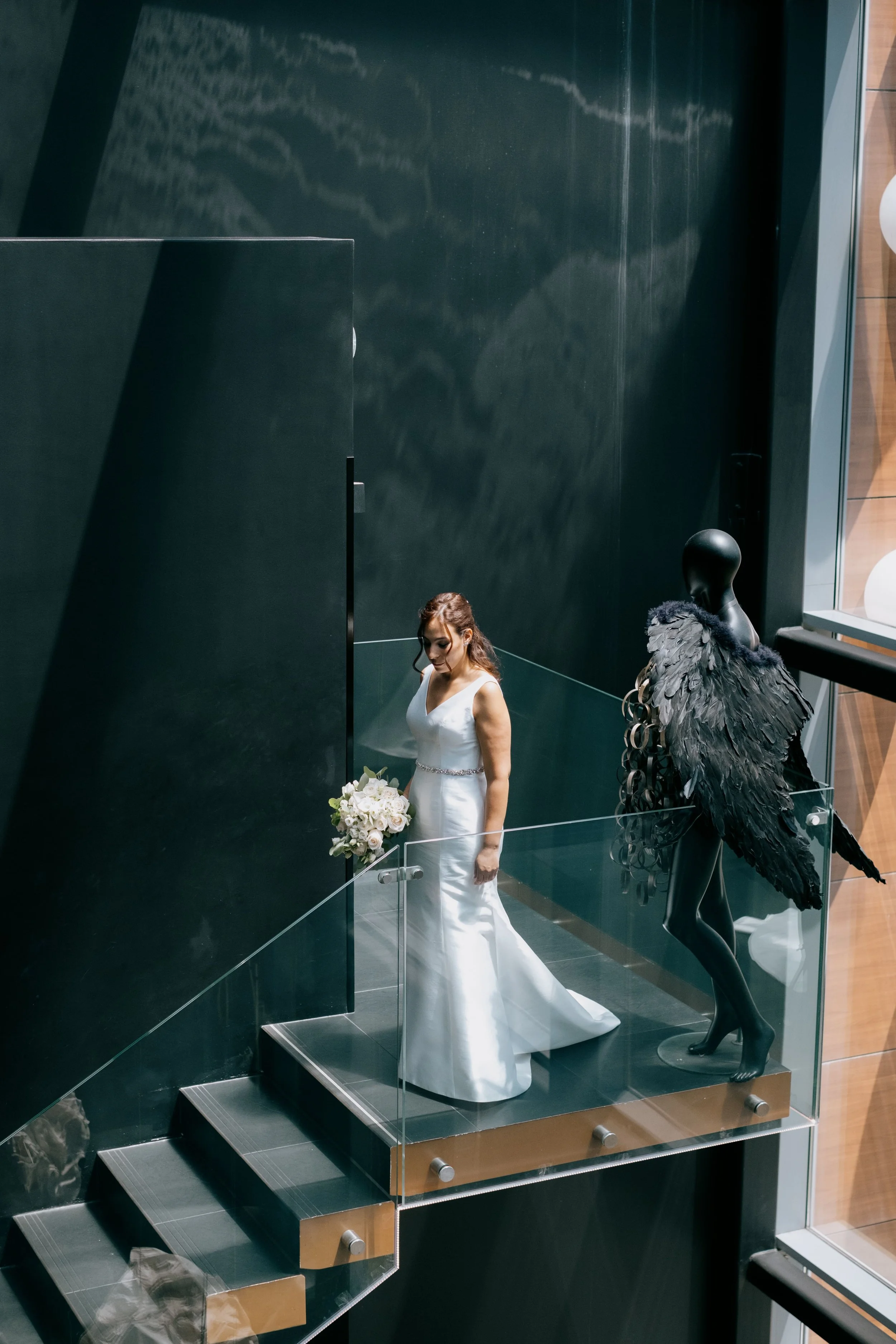 Bride in a sleek white gown descending a modern glass staircase, holding a soft pastel bouquet, beside a dark sculptural mannequin with feathered wings, captured by Carlos Mendes Photography.