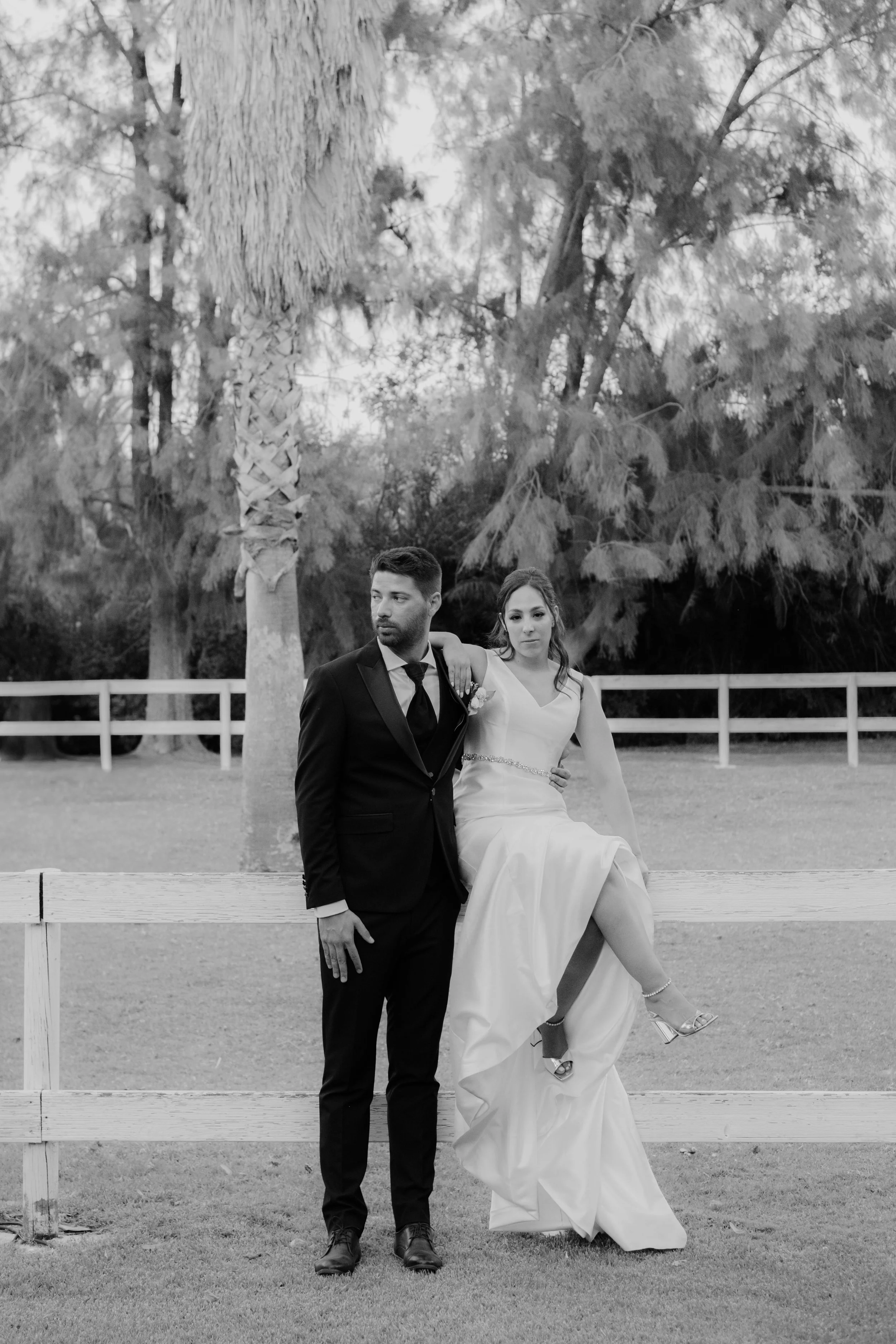 Black and white editorial-style portrait of a bride sitting on a white wooden fence with her groom standing beside her in a classic tuxedo, set in a rustic outdoor location with palm trees, captured by Carlos Mendes Photography.
