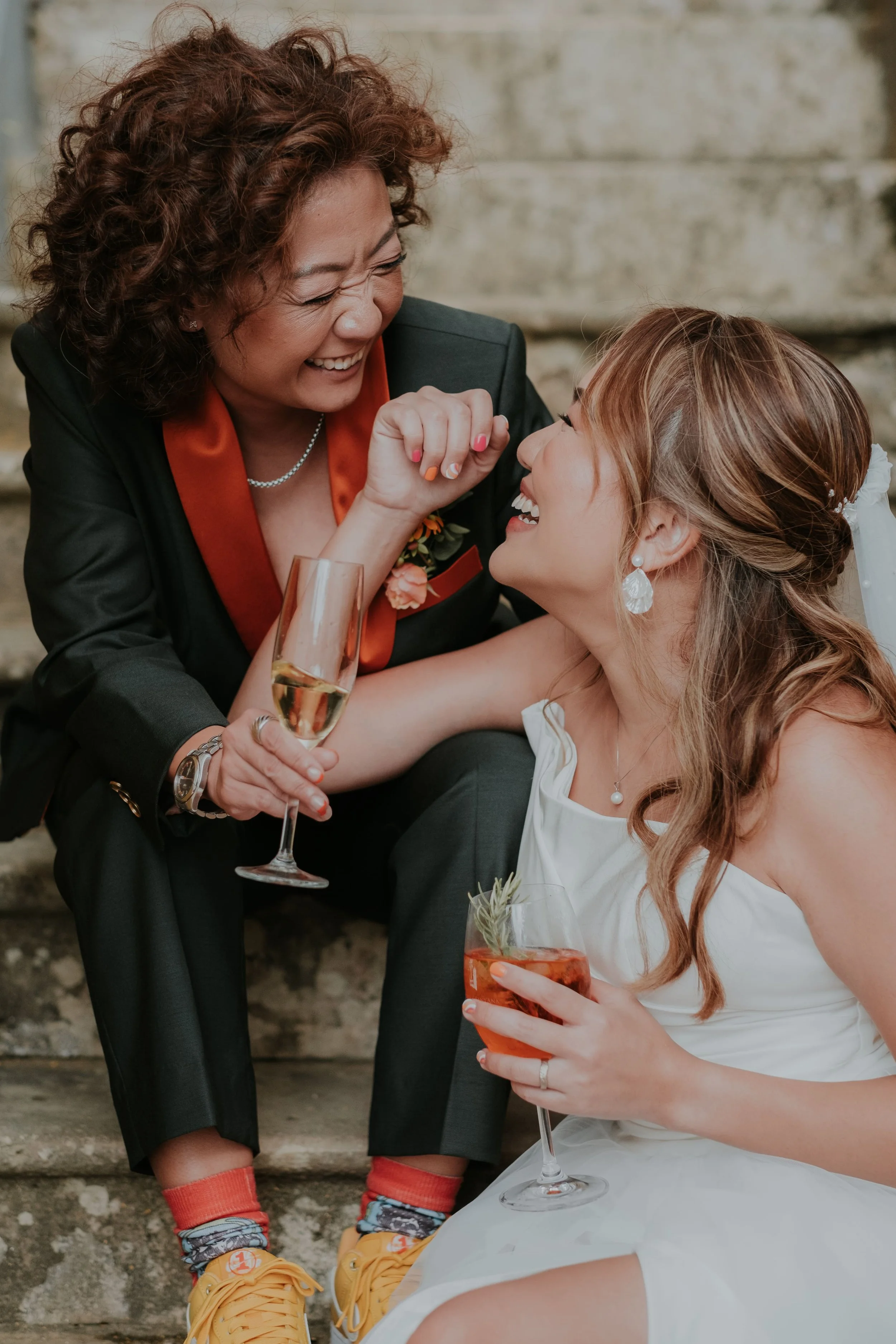 Candid moment of two brides laughing together while sitting on stone steps, holding cocktails and champagne, during their same-sex wedding celebration, captured by Carlos Mendes Photography.