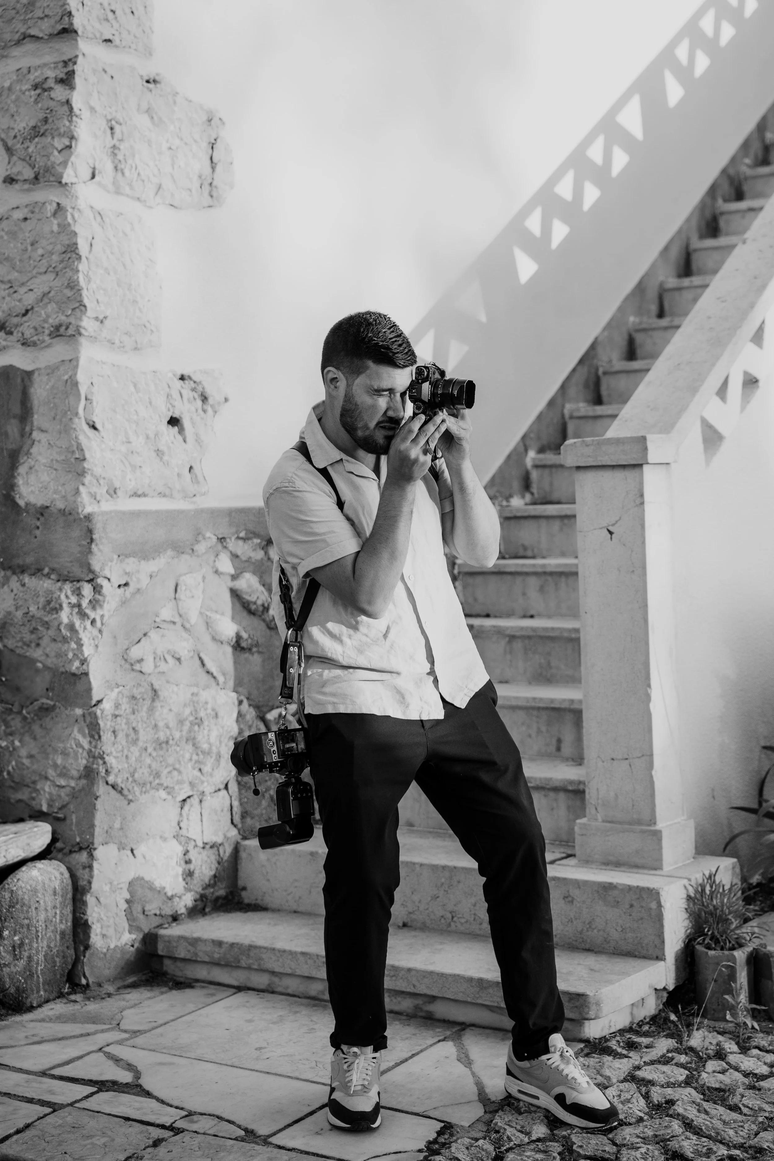 Black and white photo of professional photographer Carlos Mendes photographing with two cameras, standing in front of a rustic staircase and stone wall, captured in a candid behind-the-scenes moment.