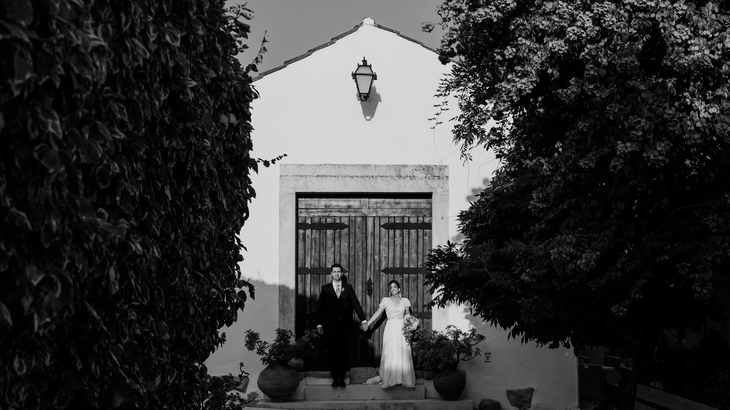 Black and white photograph of a bride and groom standing hand in hand in front of a rustic wooden door, framed by lush greenery, capturing an intimate and timeless wedding moment.