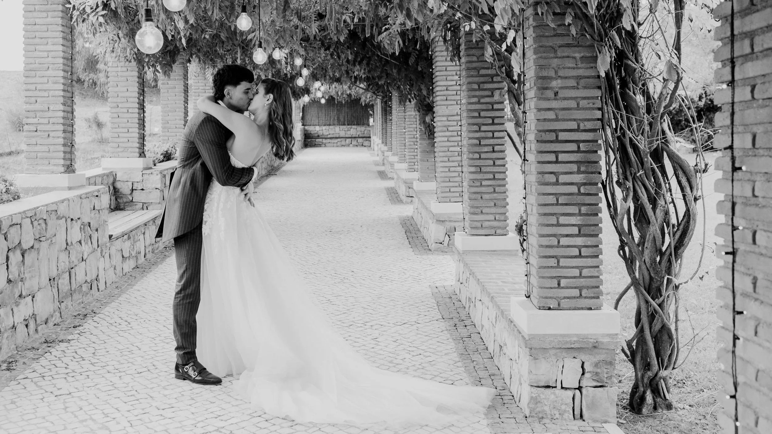 Bride and groom kissing under a vine-covered pergola with hanging lights in a black and white wedding photo.