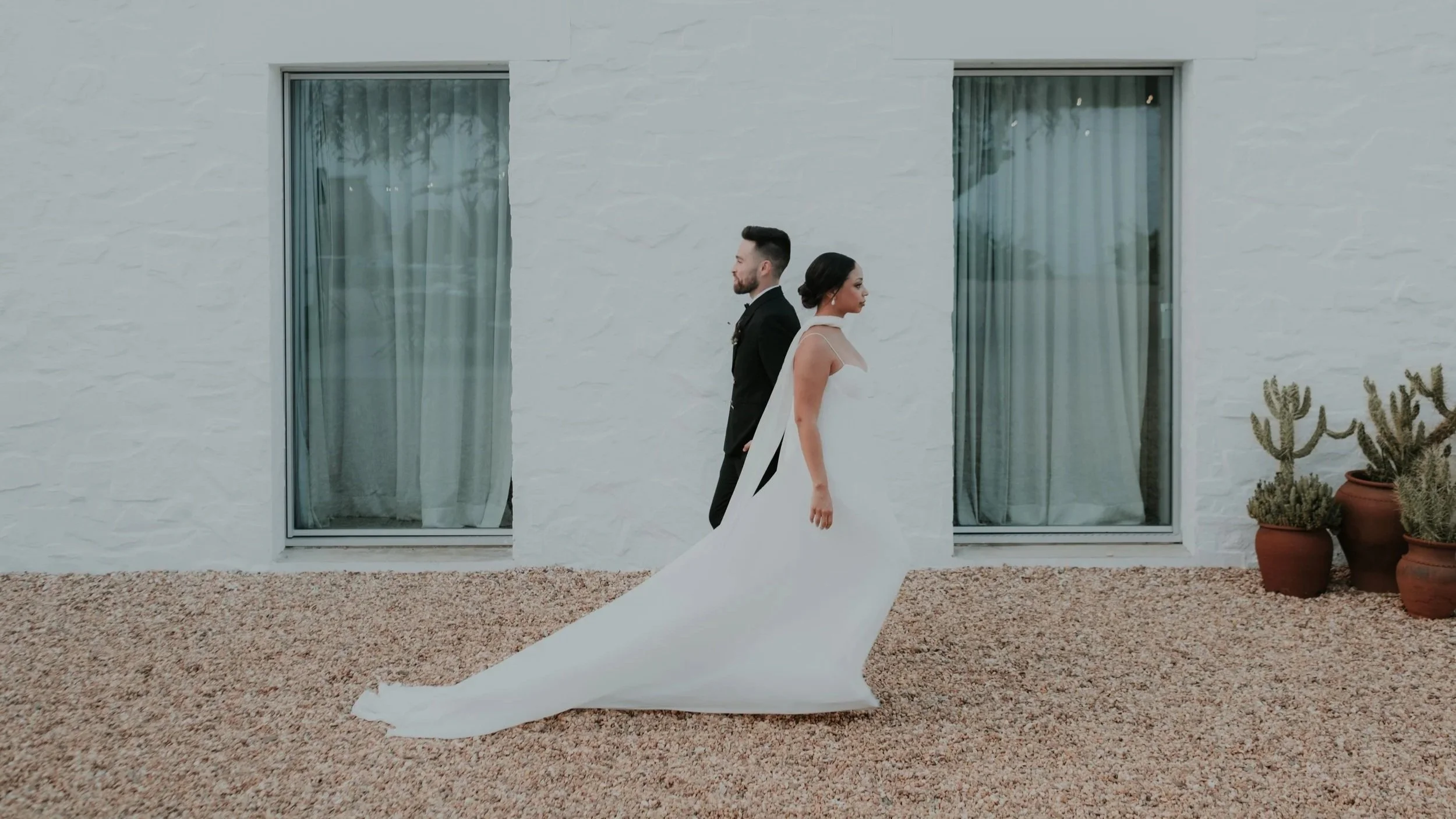 Modern wedding portrait of a bride and groom standing back to back in front of a minimalist white building with large windows and soft neutral tones, creating a clean and elegant aesthetic.