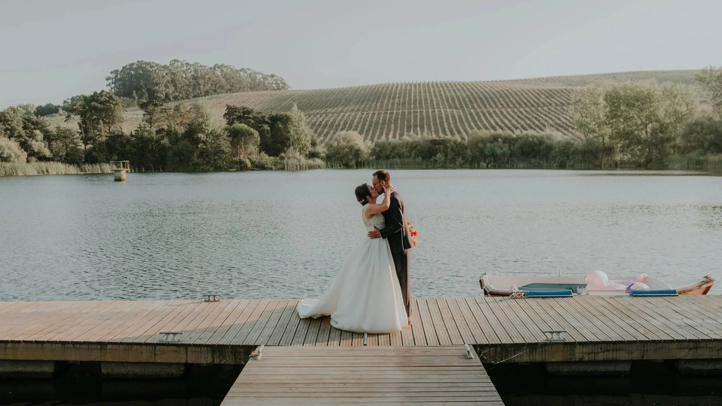 Bride and groom kissing on a wooden dock by a lake with vineyard hills in the background.