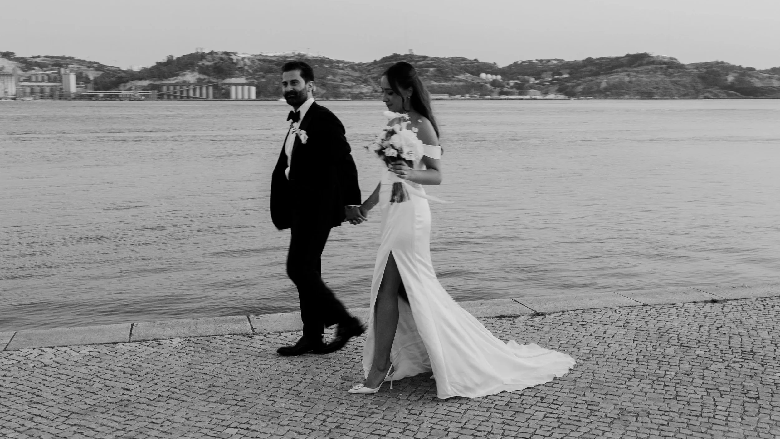 Black and white photograph of a bride and groom walking hand in hand along a waterfront promenade, with calm water and distant hills creating a timeless and elegant wedding atmosphere.
