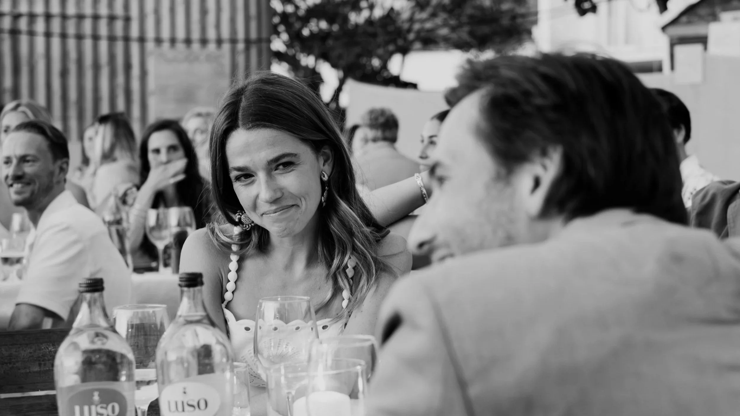 Woman smiling at a table during a wedding reception with guests and drinks in a black and white photo.