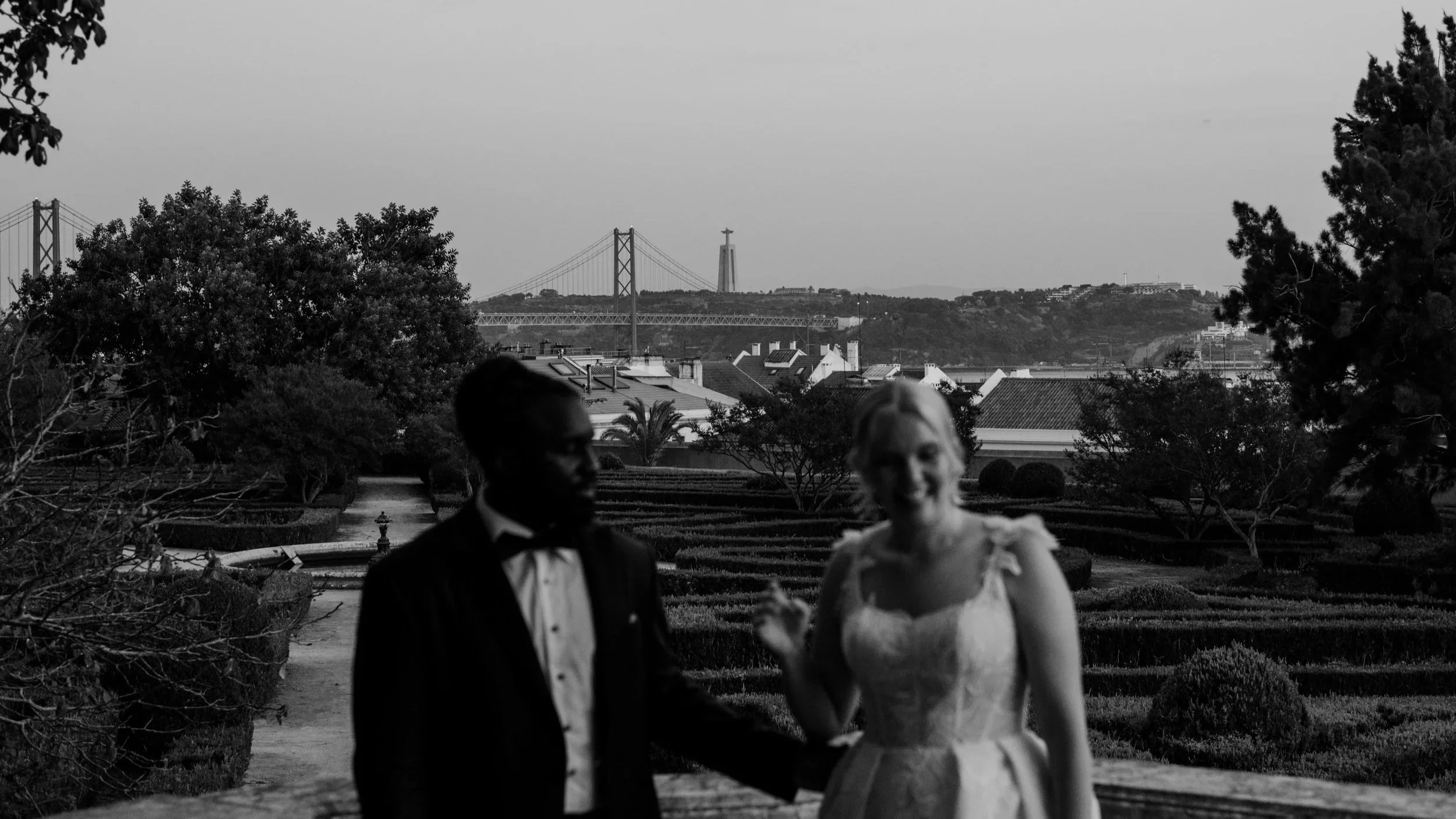 Black and white photograph of a bride and groom standing in a landscaped garden overlooking a city skyline, with a suspension bridge and monument in the distance, creating a romantic and scenic wedding atmosphere.