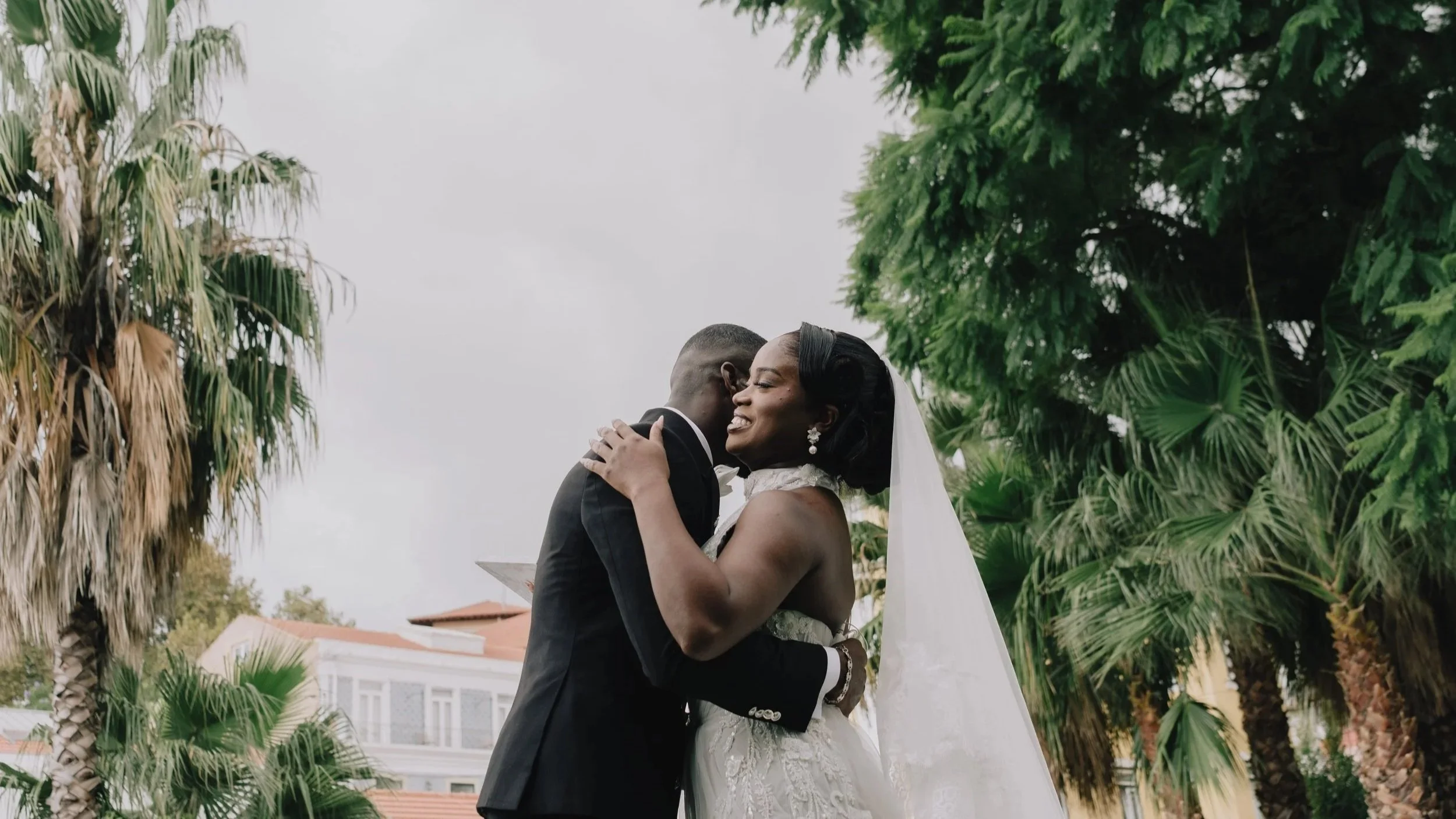 Romantic wedding moment of a bride and groom embracing outdoors, surrounded by lush palm trees and greenery, creating a warm and elegant destination wedding atmosphere.