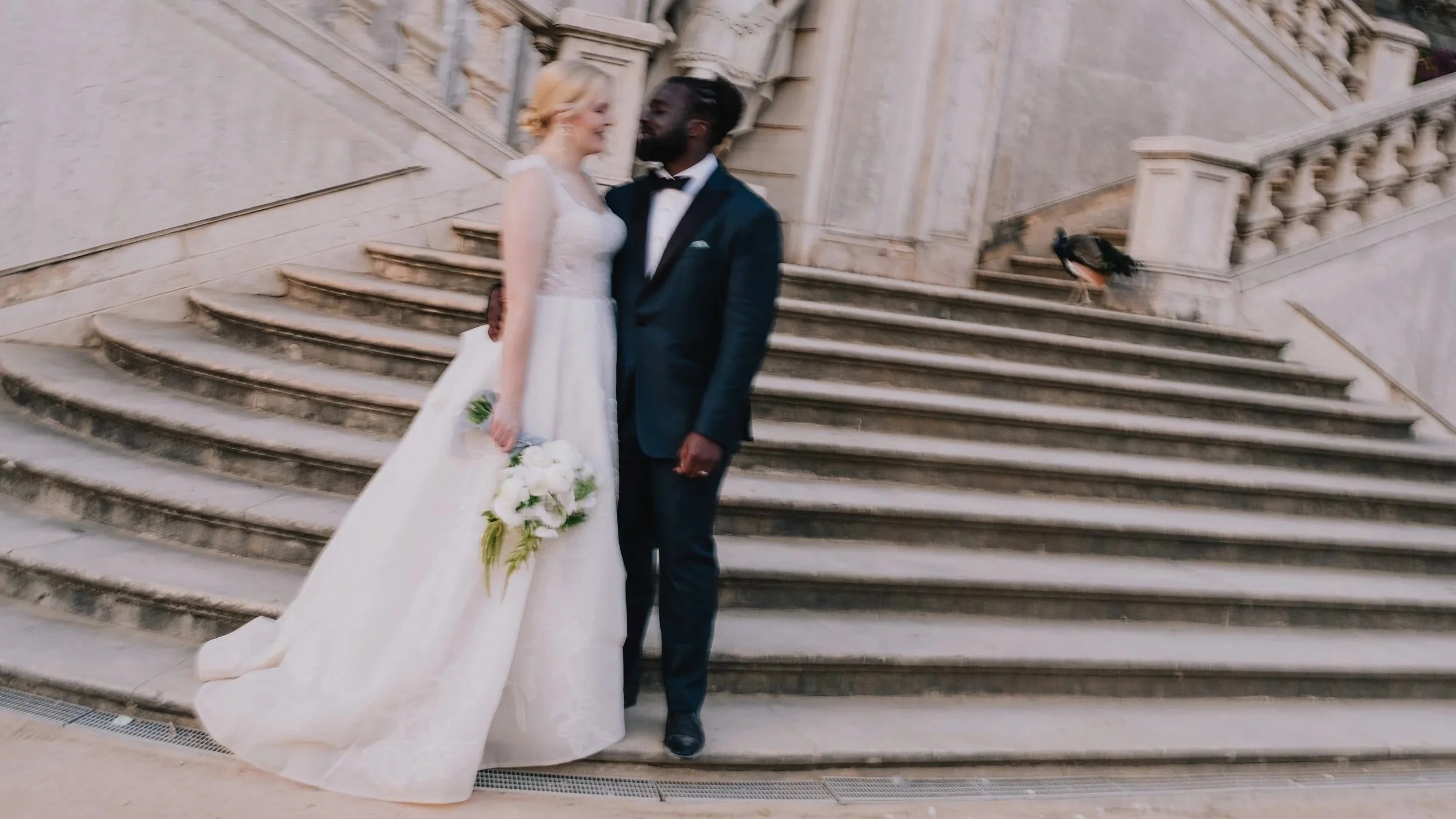 Romantic wedding portrait of a bride and groom standing on grand stone stairs in front of a classical architectural building with statues, creating a refined and timeless atmosphere.