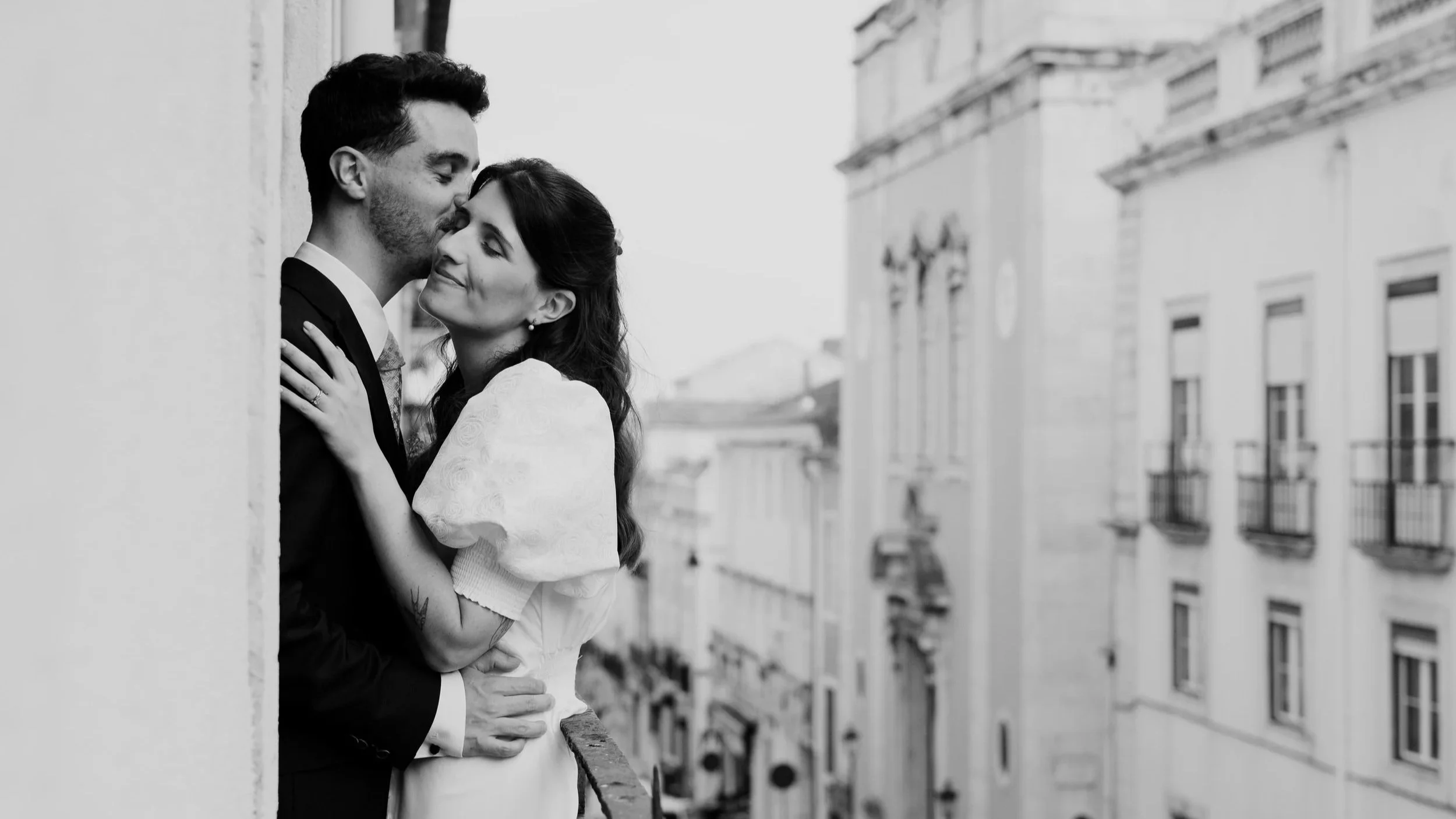 Black and white photograph of a bride and groom embracing on a balcony overlooking a historic European city street, capturing a romantic and intimate wedding moment.