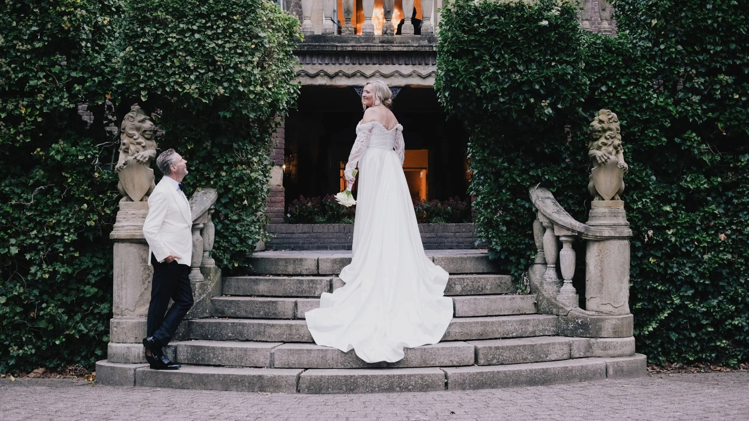 Elegant wedding scene of a bride standing on grand stone steps outside a historic building covered in greenery, while the groom looks up at her, creating a romantic and timeless atmosphere.
