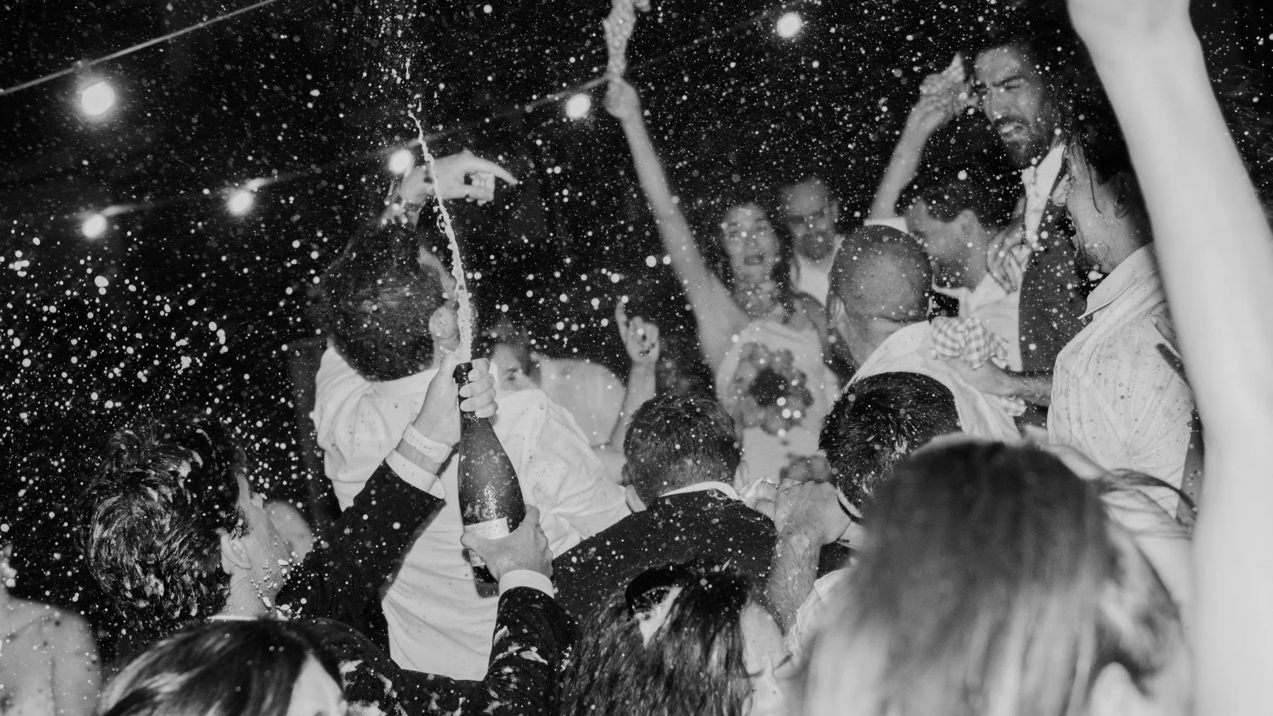 Pessoas celebrando uma festa com muita espuma, algumas segurando garrafas de bebida, em um ambiente escuro com luzes.