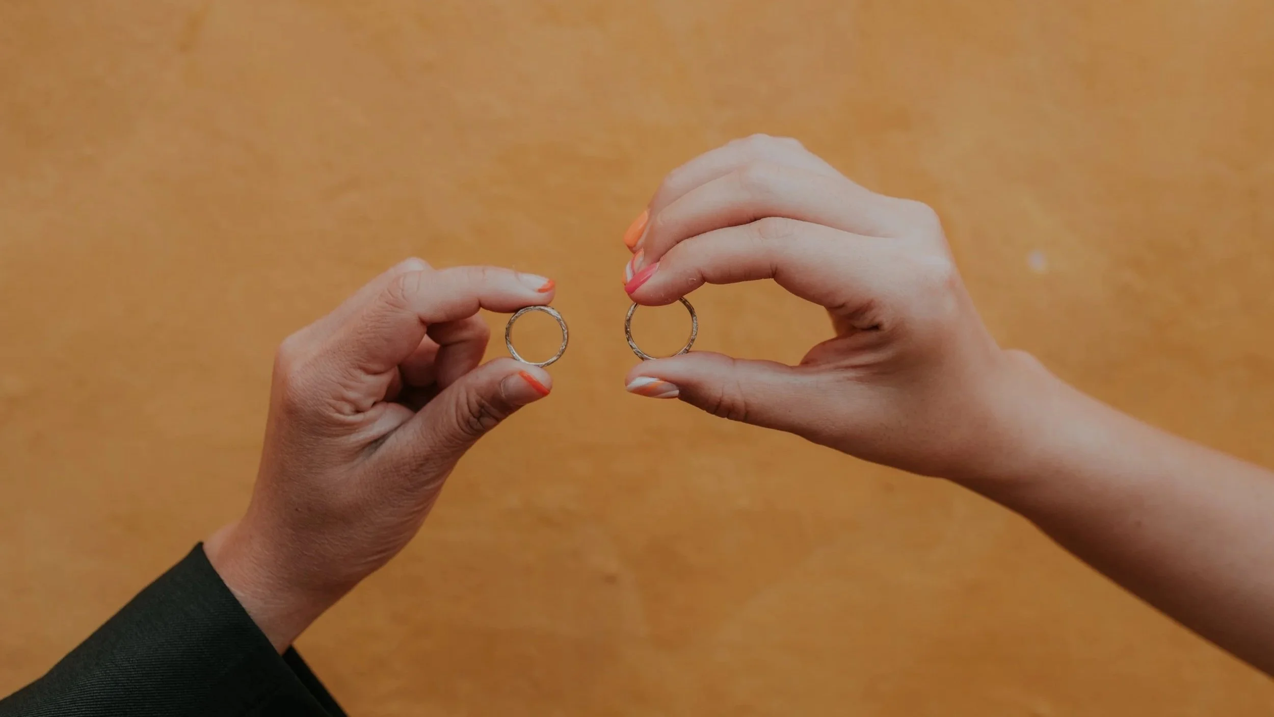 Close-up of two hands holding minimalist metal rings against a warm earthy background, representing connection, unity, or commitment in a modern aesthetic.