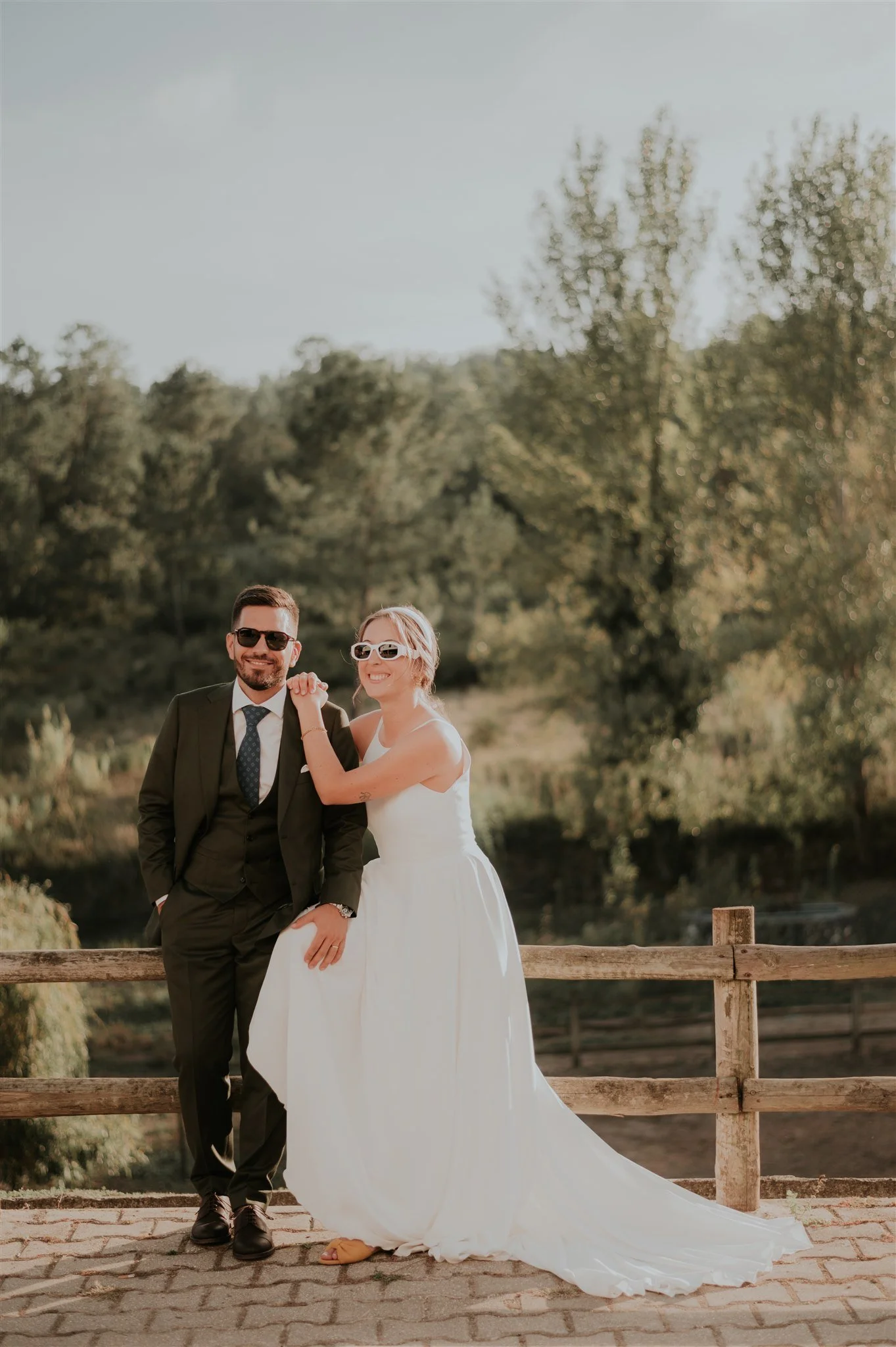 Casal de casamento em pose descontraída ao ar livre, com árvores verdes ao fundo e uma cerca de madeira na frente, na luz do pôr do sol.