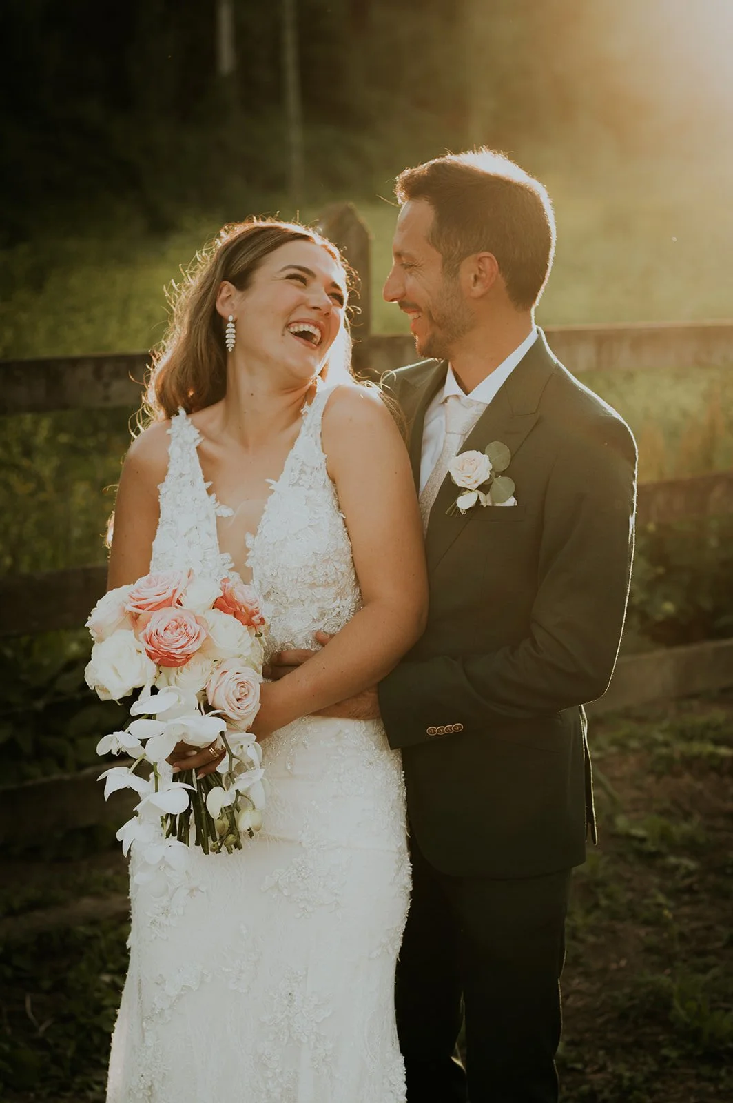 Casal de noivos sorridente ao ar livre durante o pôr do sol, com a mulher segurando um buquê de rosas e orquídeas brancas, ambos vestidos com trajes de casamento.