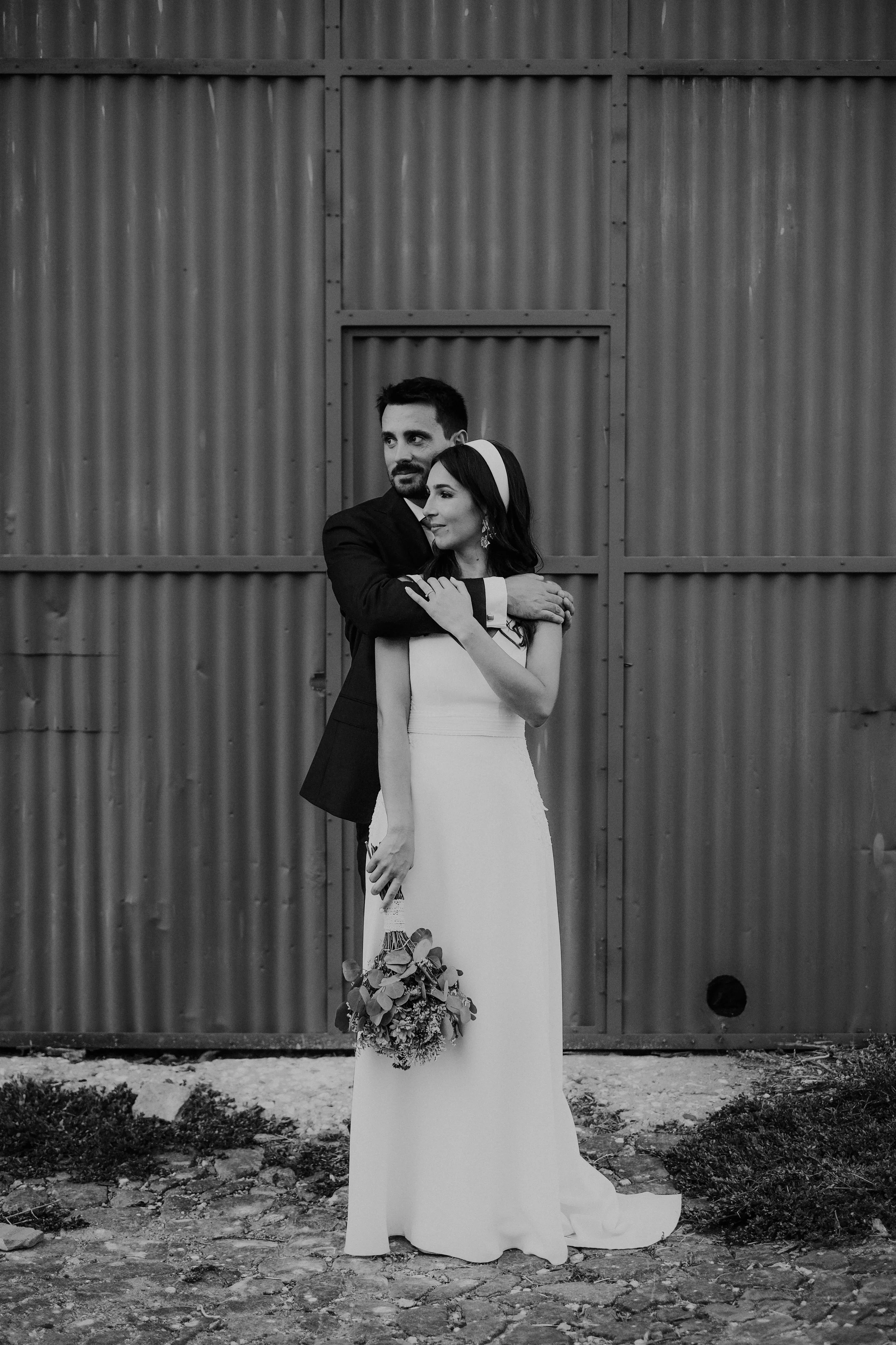 Casal de noivos posando lado a lado, com o homem abraçando a mulher pelos ombros, em frente a uma parede de metal. A mulher segura um buquê de flores e usa vestido branco e lenço na cabeça. A imagem é em preto e branco.