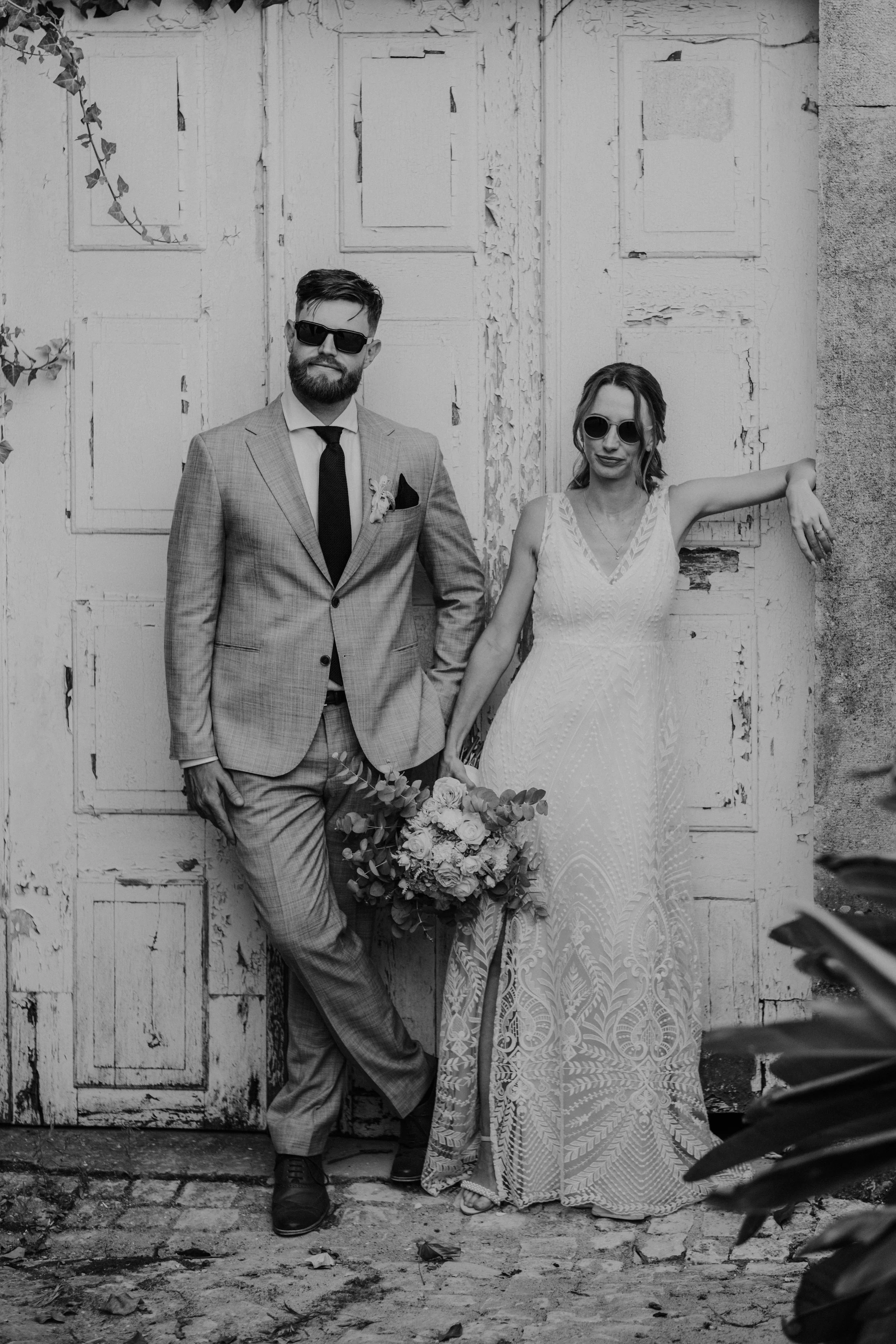 Casal de noivos com roupas elegantes, ele de terno e ela de vestido, em frente a uma porta antiga e desgastada, ambos usando óculos escuros, ela segurando um buquê de flores.