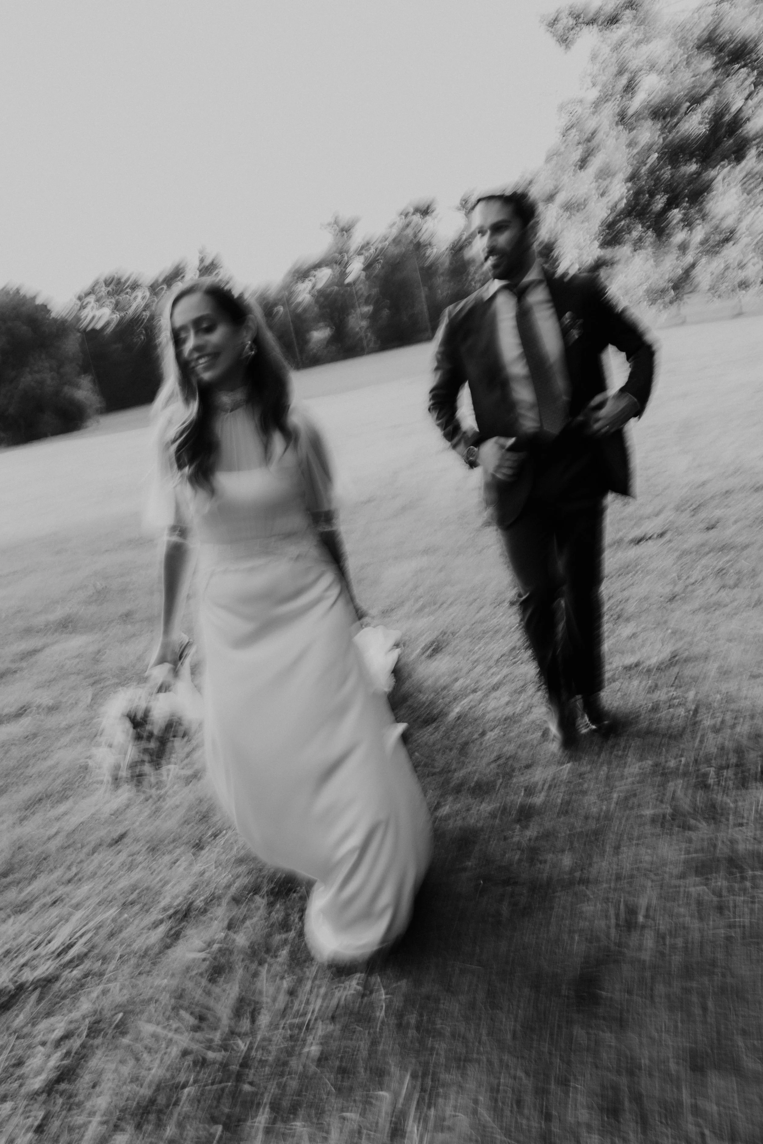 Imagem em preto e branco de um casal andando na grama, com árvores ao fundo, aparentemente em um parque, vestido formalmente, possivelmente em um casamento ao ar livre.