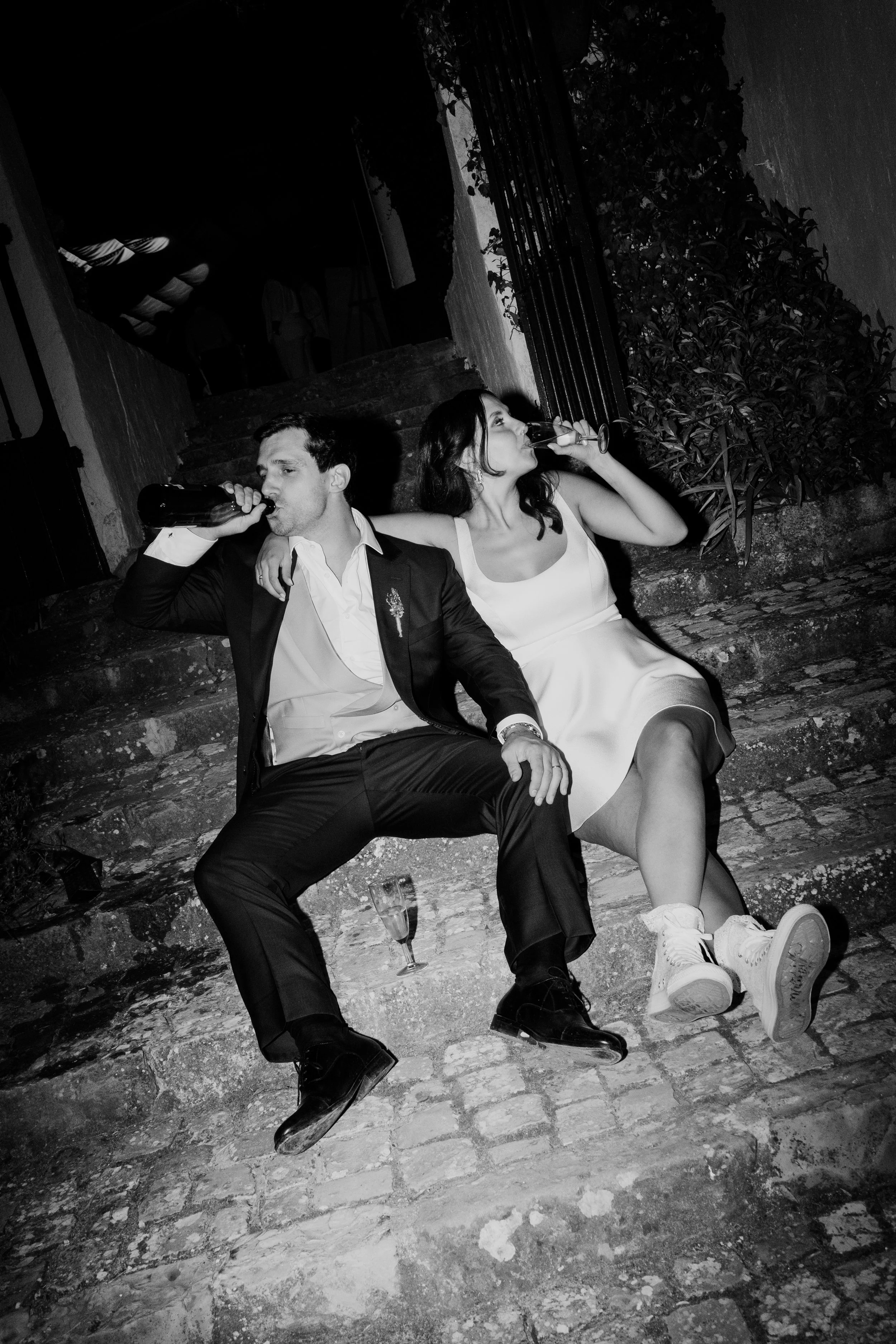 Homem e mulher sentados nos degraus de uma escadaria, ambos bebendo de garrafas, com roupa formal, em ambiente à noite, ao ar livre, em preto e branco.