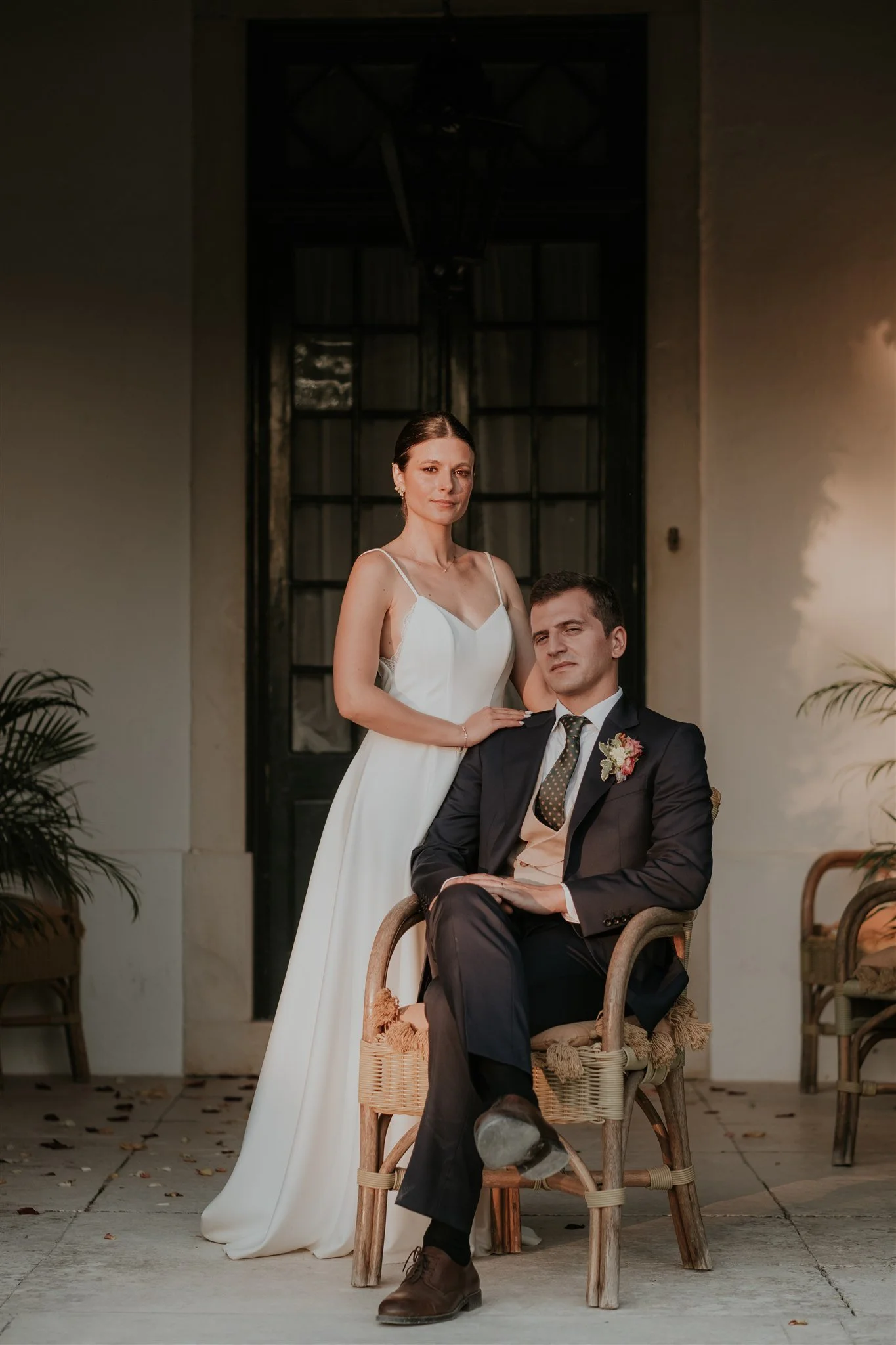 Casal de casamento posando para foto, a mulher usando vestido branco e o homem de terno, sentados e de pé, em ambiente externo com plantas e cadeiras de vime.