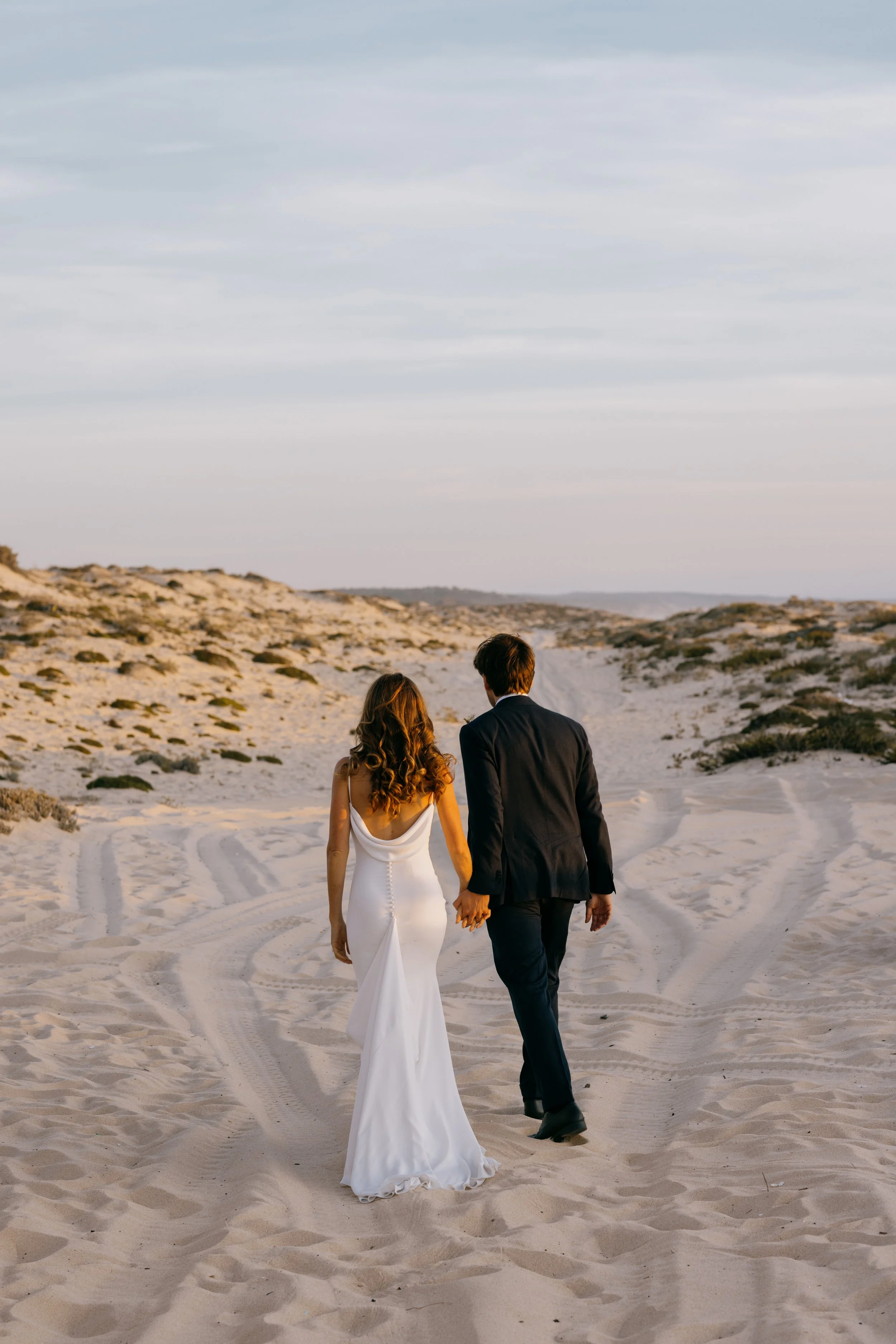 Casal de vestido de casamento e terno deitado na praia, caminhando de mãos dadas durante o pôr do sol.