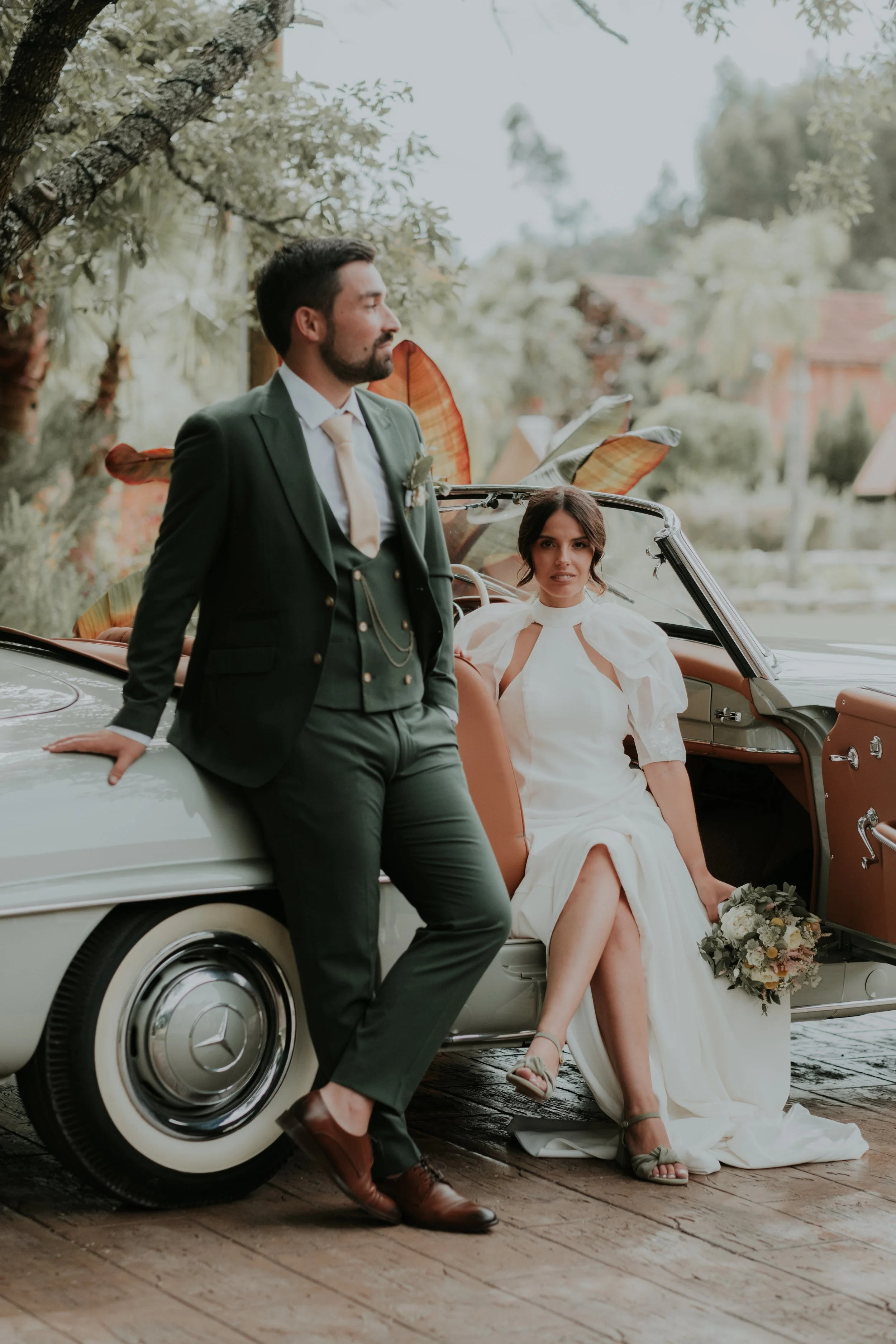 Casal em casamento com carro vintage ao ar livre, mulher de vestido branco com buquê, homem de terno verde escuro, cenário natural.