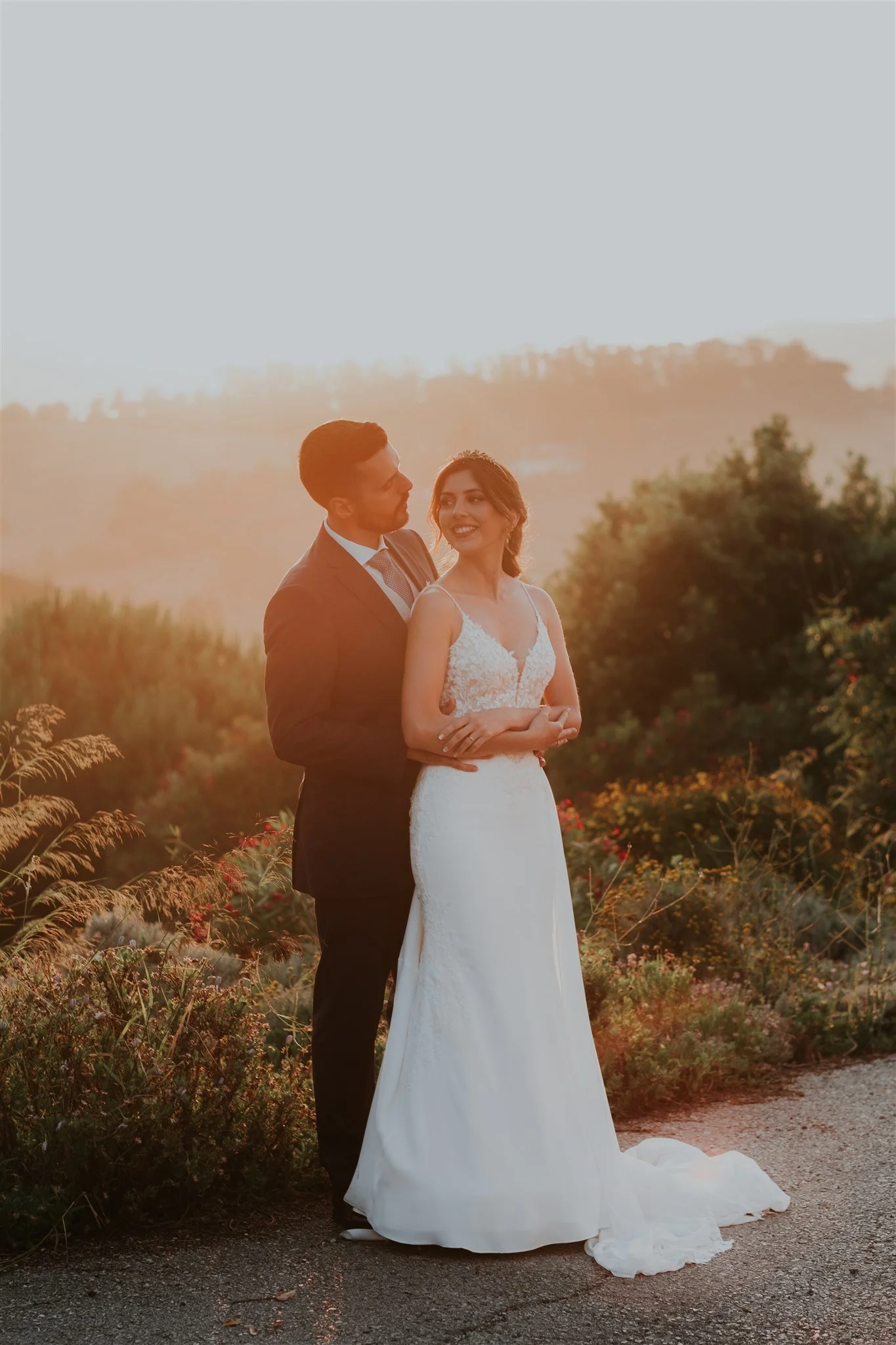 Casal de noivos de casamento ao pôr do sol, com decoração natural ao ar livre, mulheres com vestido de noiva e homens com terno, sorrindo e olhando um para o outro.