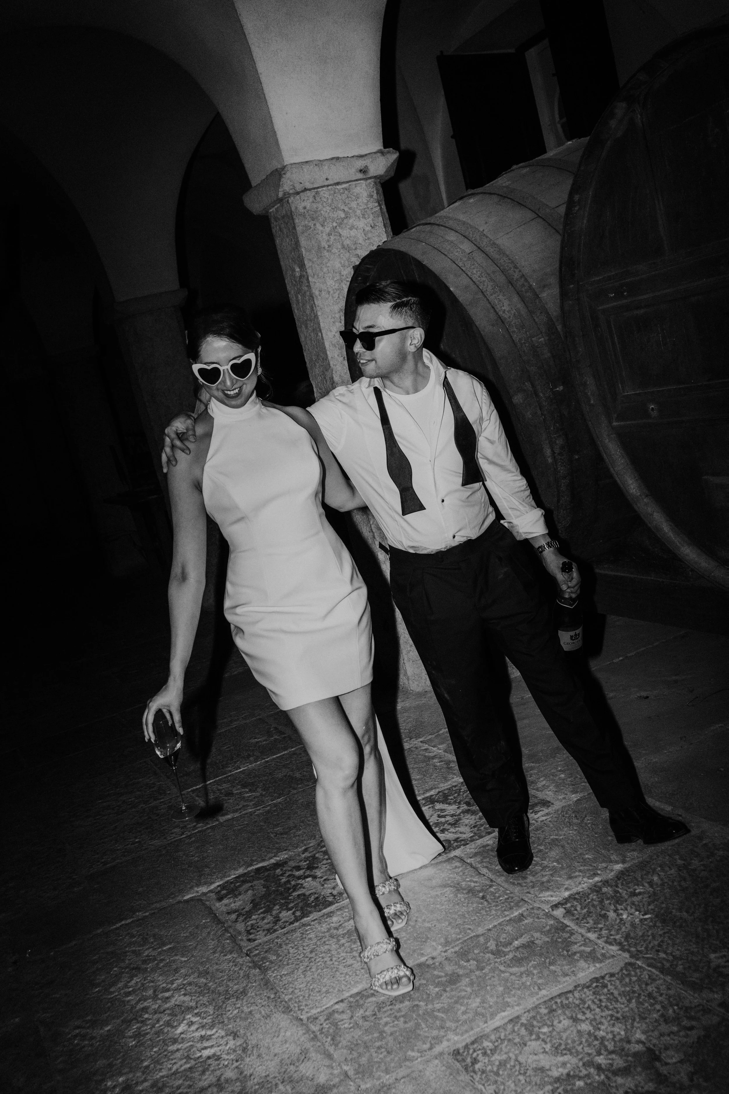 Casal celebrando com roupas elegantes, sorrindo, usando óculos de sol e brindando em um ambiente com grandes barris de madeira ao fundo, em tom preto e branco.