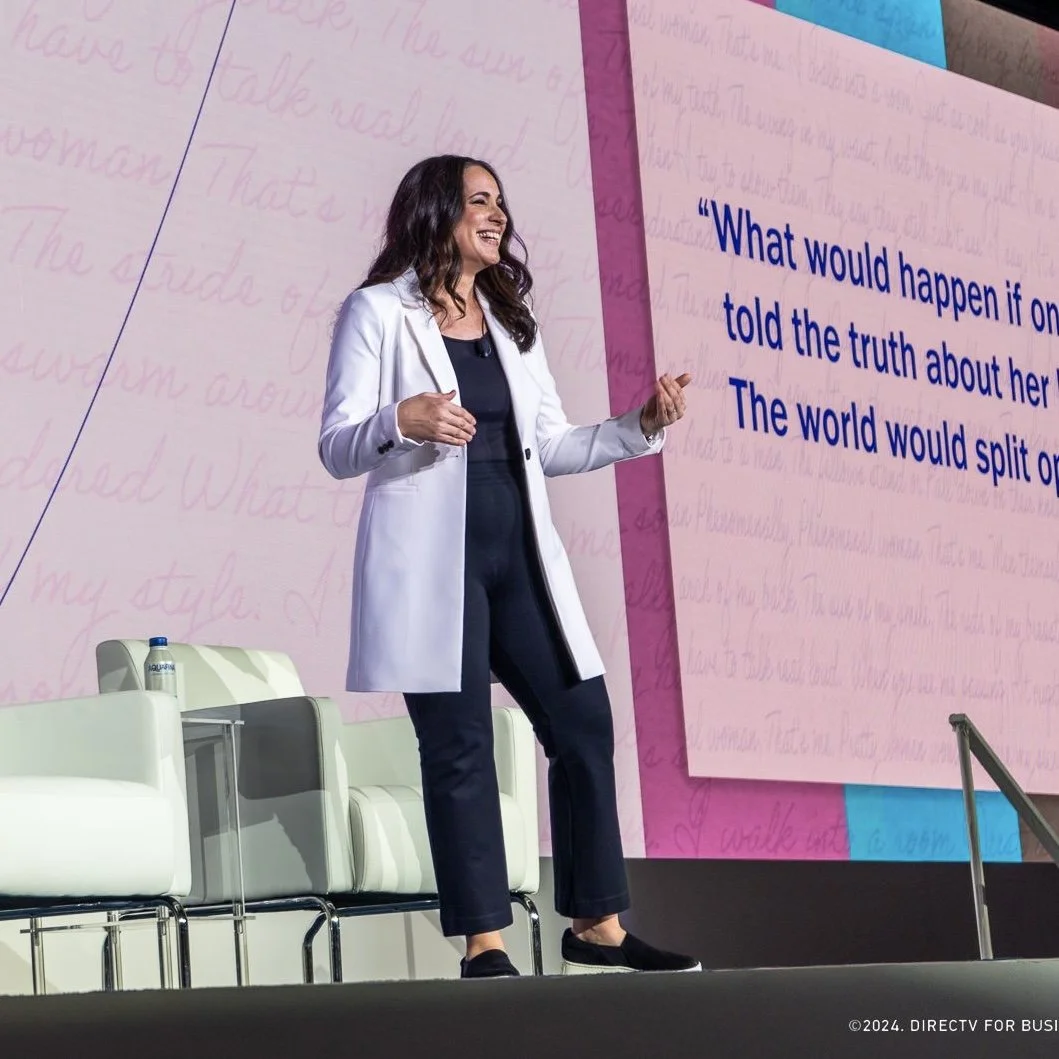 Anna Blue in a white blazer and dark pants speaking on stage with a large screen behind her displaying a quote in blue text, with chairs and a water bottle on stage.