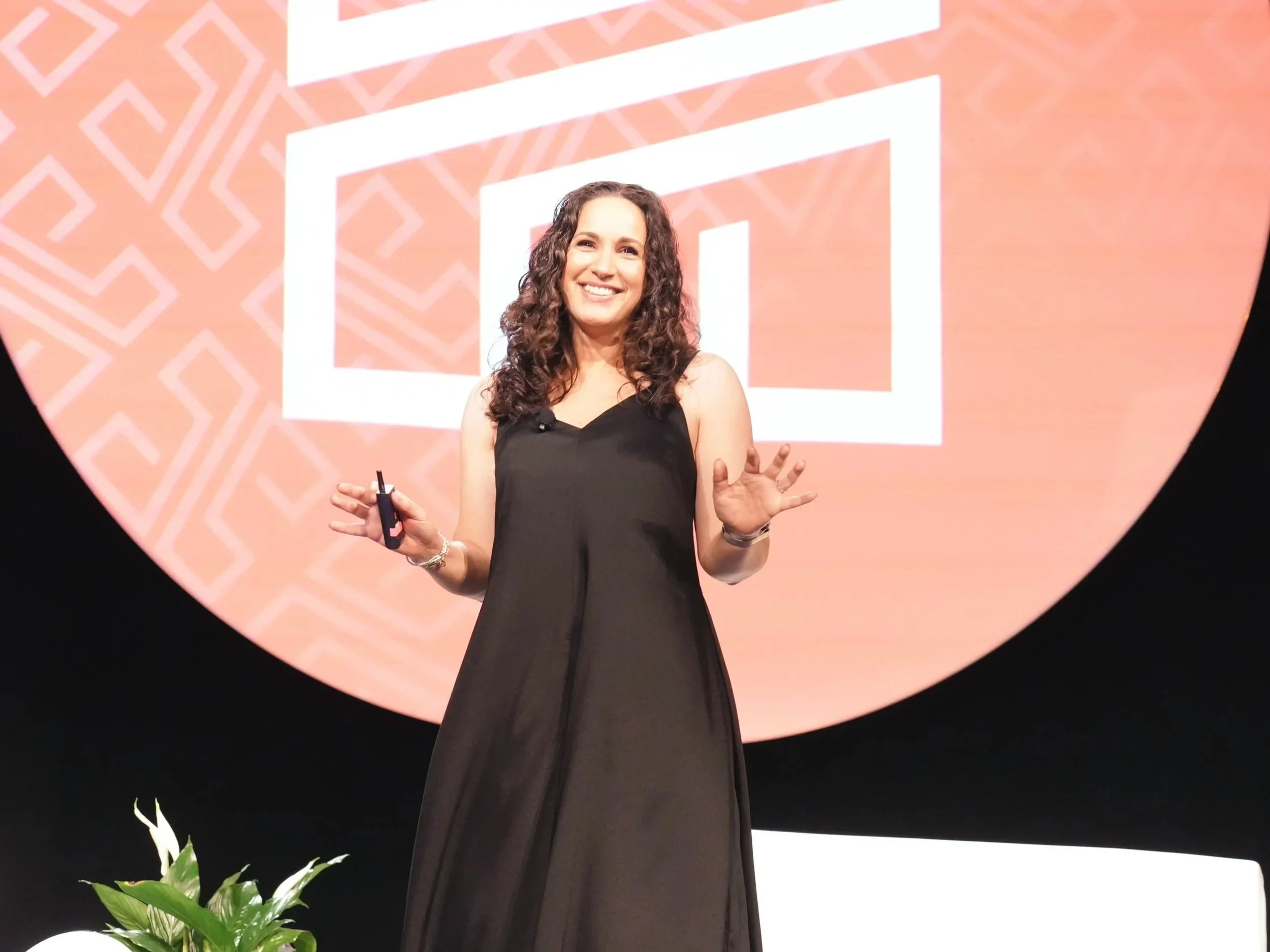 A woman smiling and speaking on stage during a presentation, holding a remote control, with a large screen behind her displaying a geometric logo in pink and white.