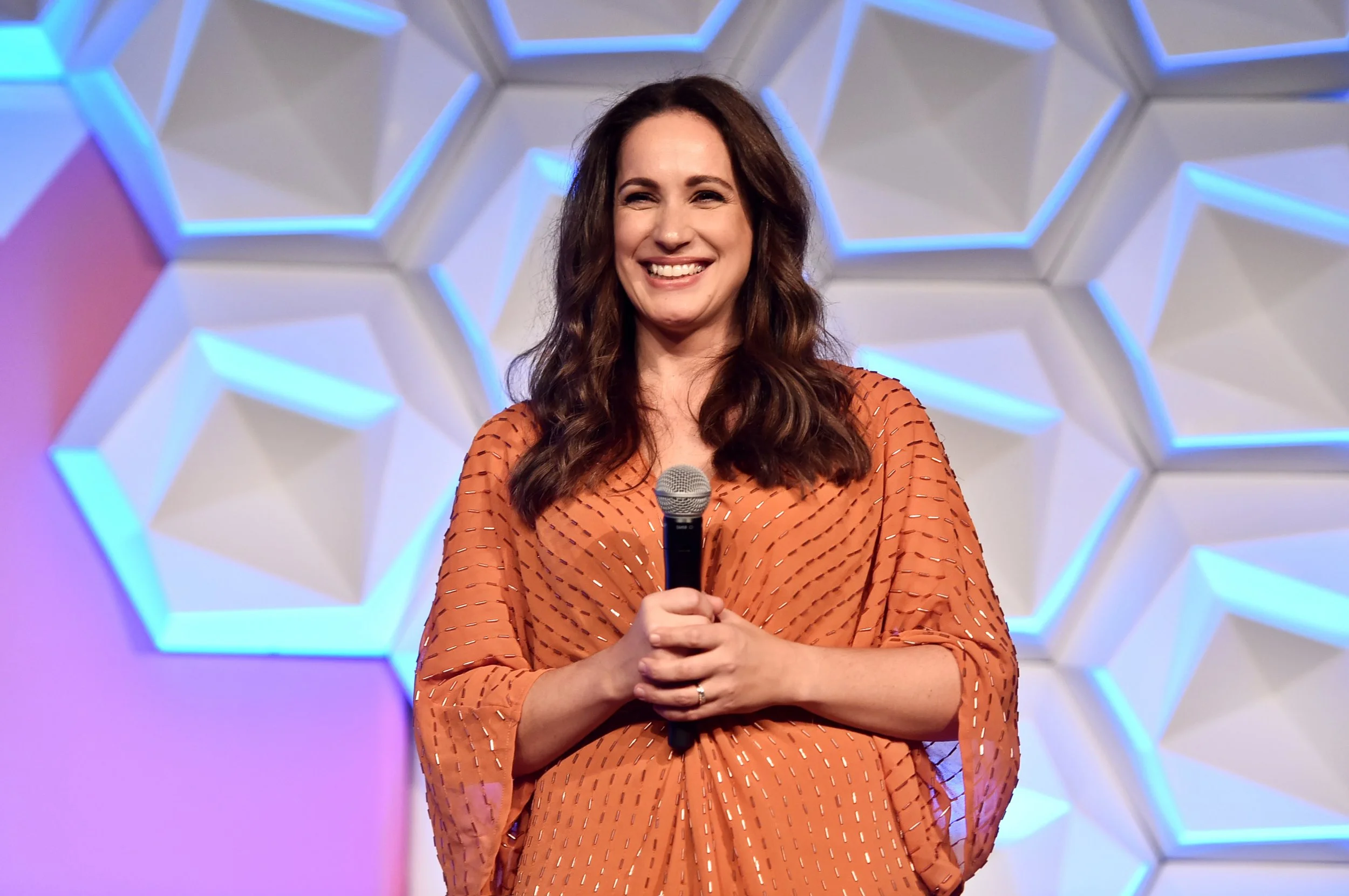 Anna Blue with long brown hair smiling and holding a microphone on stage with geometric white and blue background.