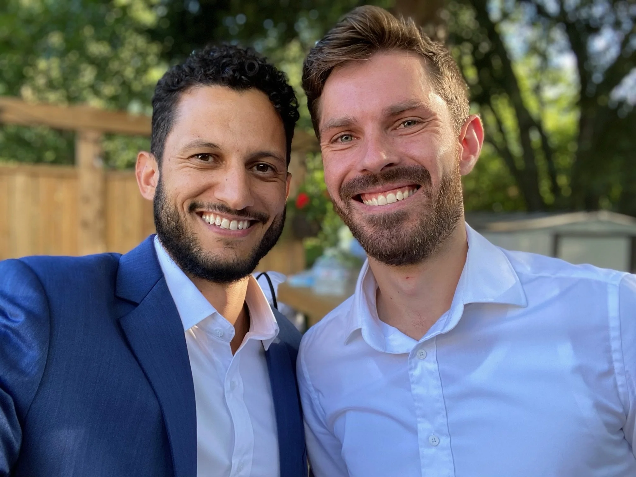 Two smiling men taking a selfie outdoors in a sunny setting with trees and a wooden fence in the background.