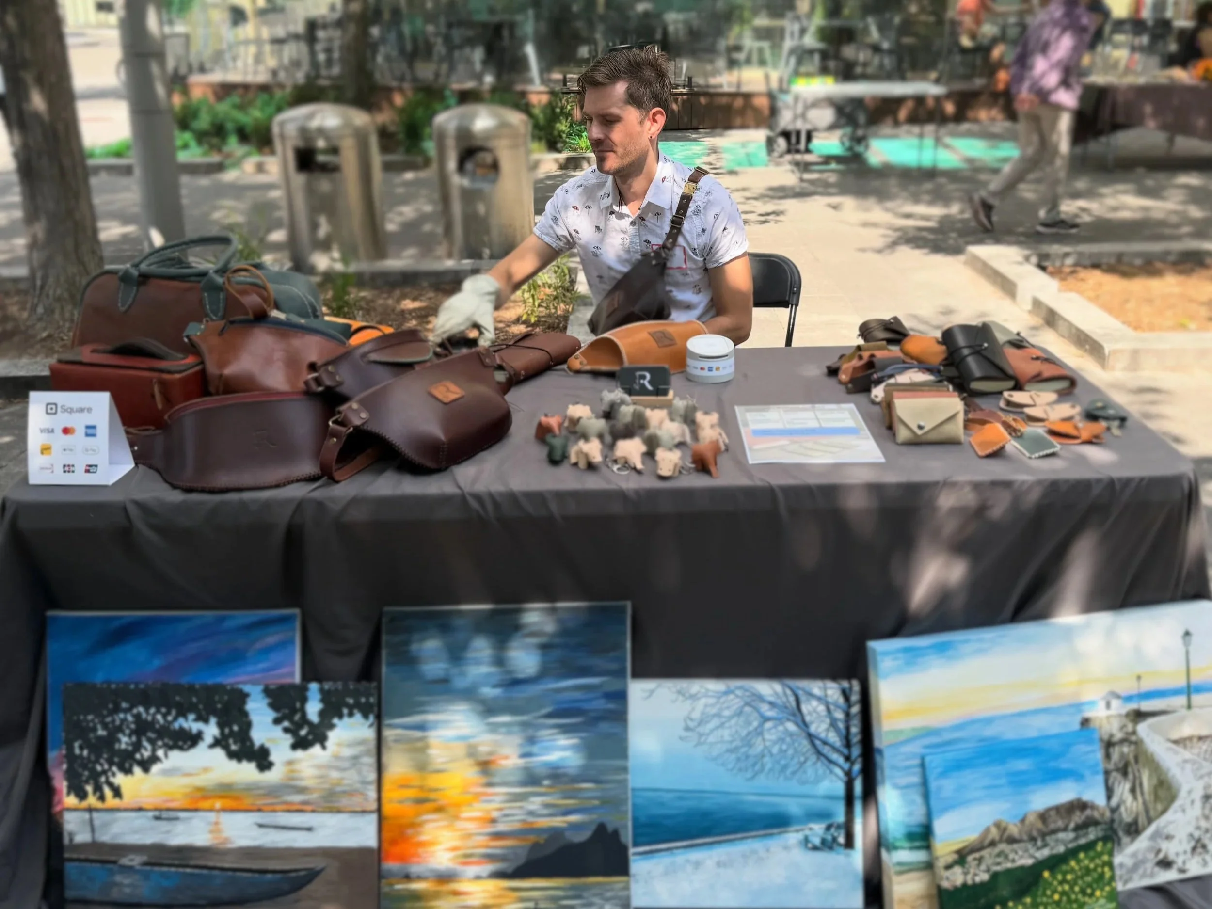 A man at an outdoor market stall displaying leather bags, wallets, and art paintings of natural scenes on a table covered with black cloth.