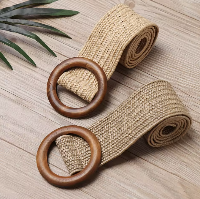 Two rolled beige fabric belts with wooden circular buckles on a wooden surface, with green leaves on the side.