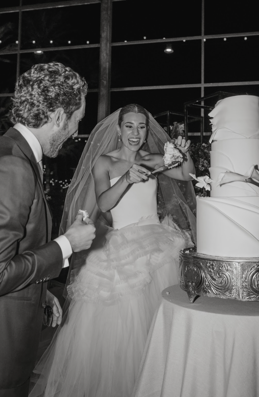 A bride and groom sharing a piece of wedding cake during their reception, with the bride smiling and holding a fork with cake, and a large wedding cake on a table beside them.