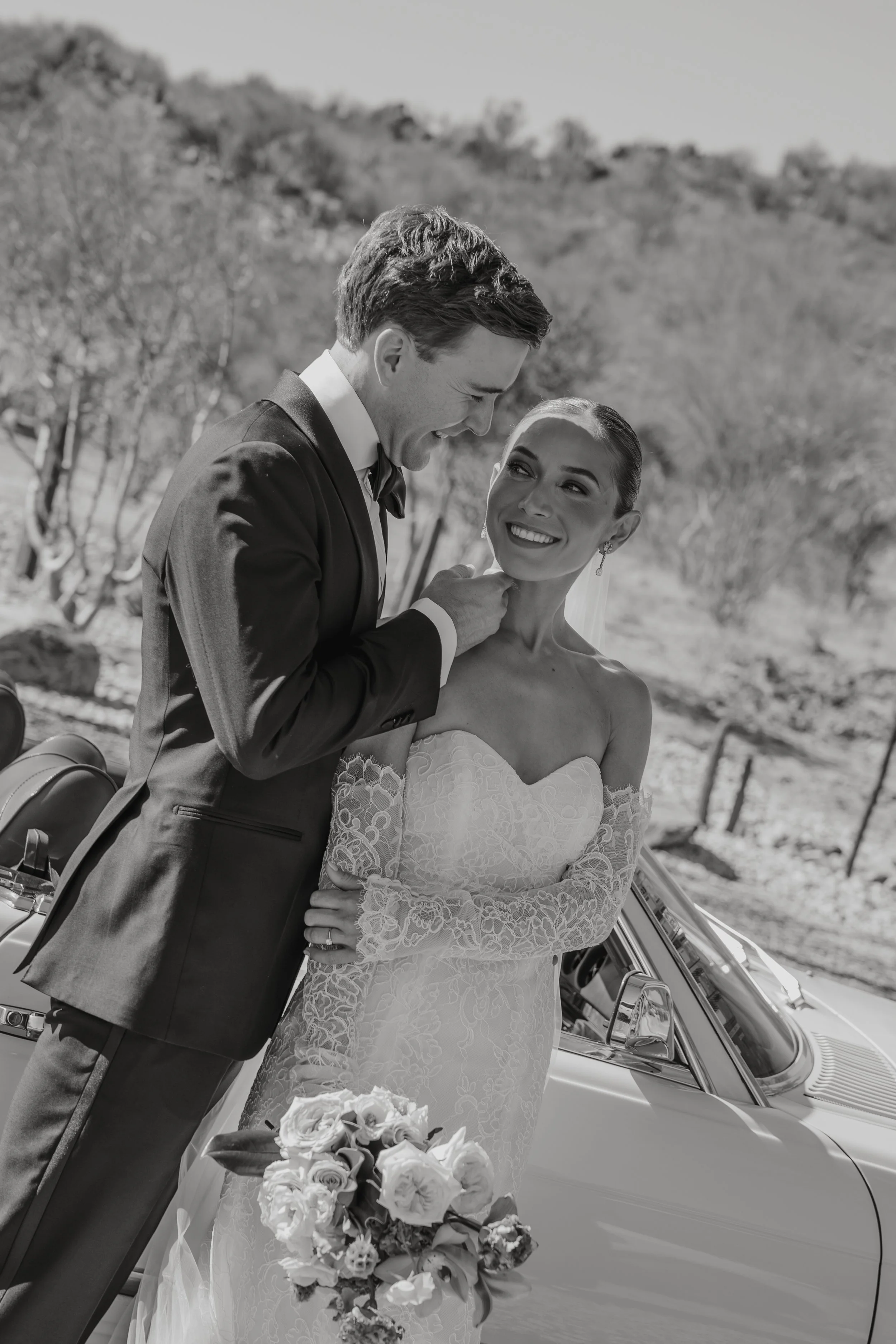 Black and white photo of a bride and groom outdoors, with the groom smiling and touching the bride's face, and the bride smiling back holding a bouquet of flowers, next to a classic car.